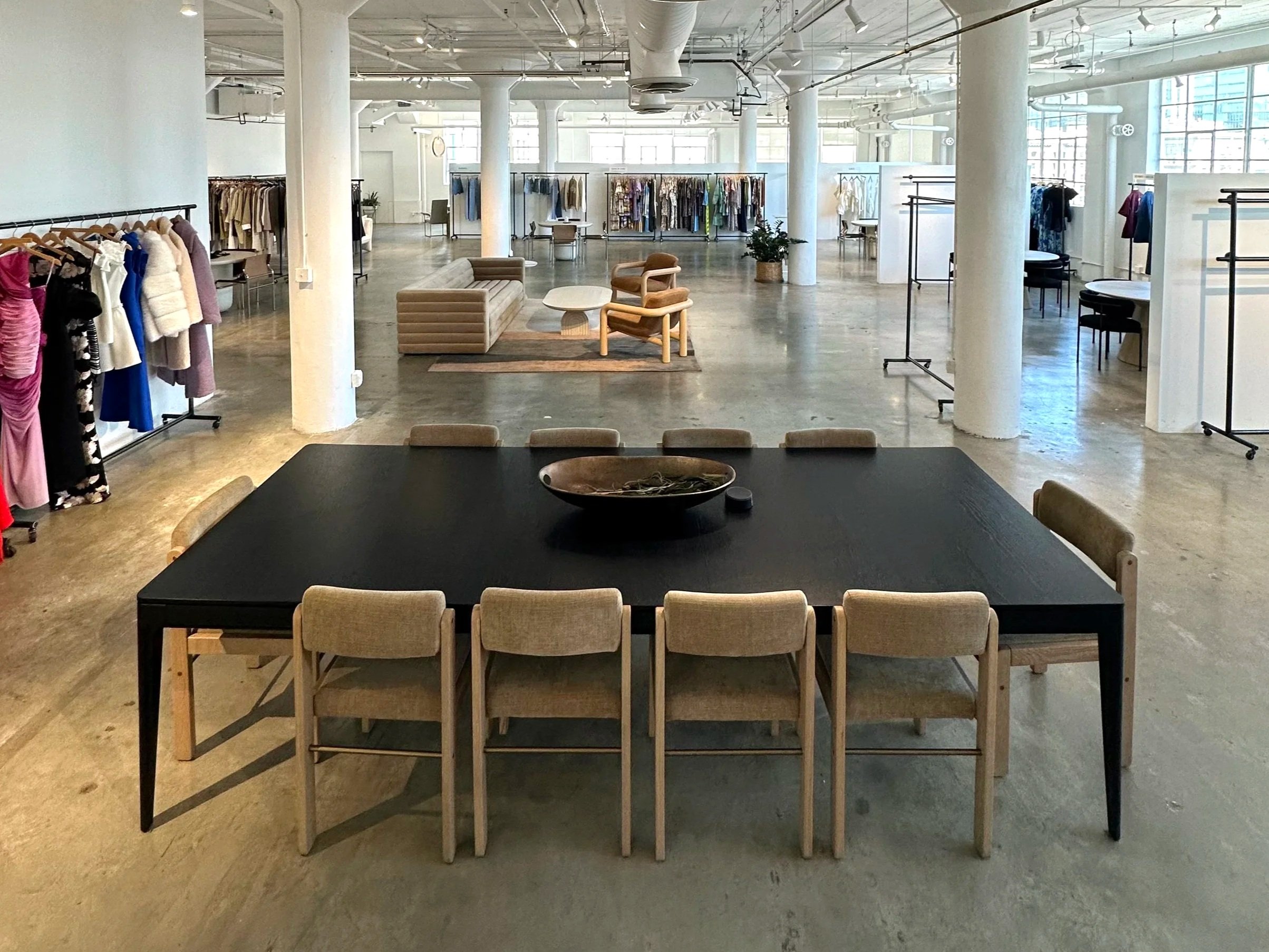 Interior of a clothing store with racks of clothes, a sitting area with chairs and a table, and a black dining table with beige chairs in the foreground. The space features white columns, large windows, and industrial-style ceiling with exposed pipes