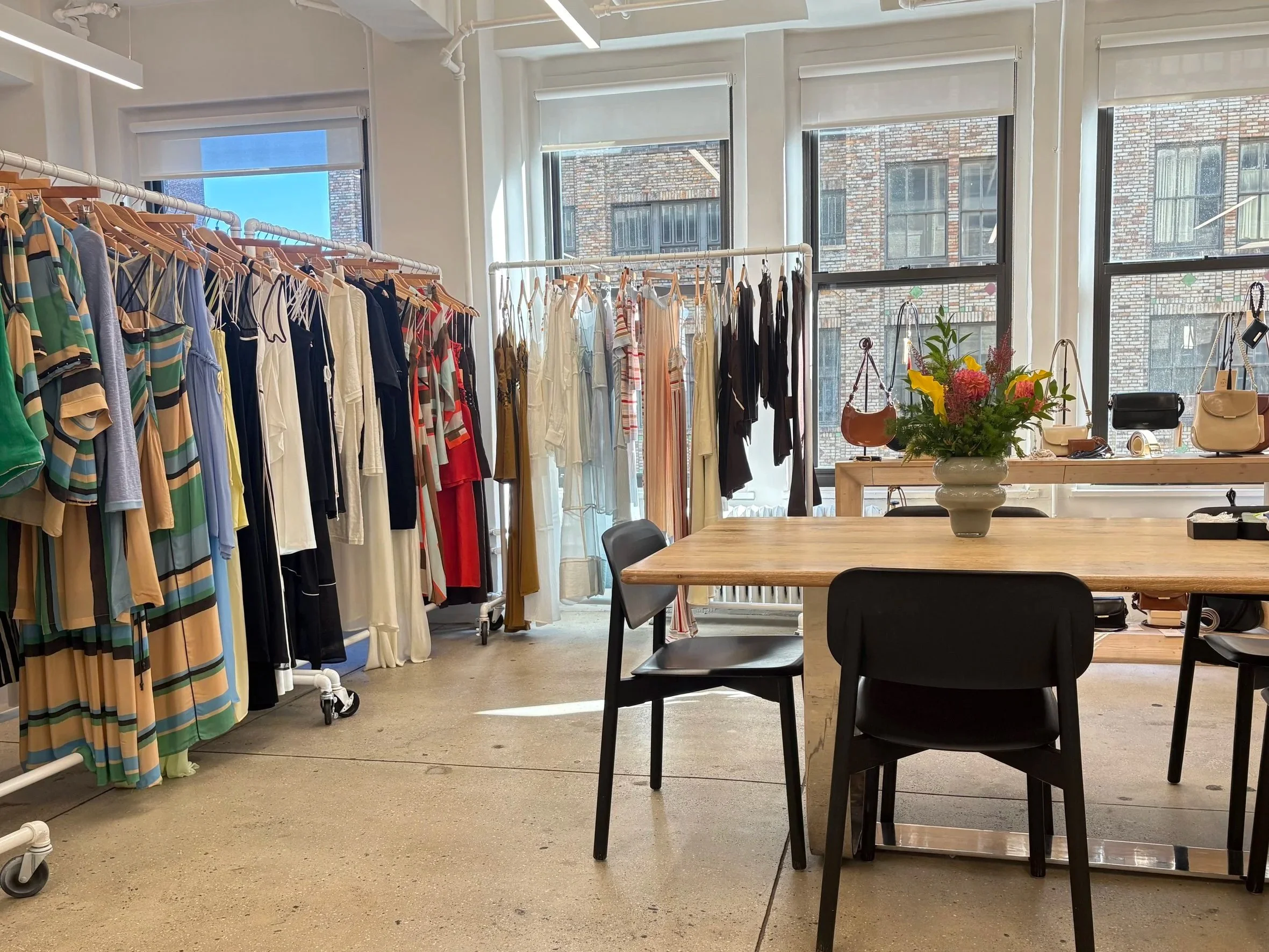 Clothing store interior with racks of dresses, handbags on a table, and large windows letting in natural light.