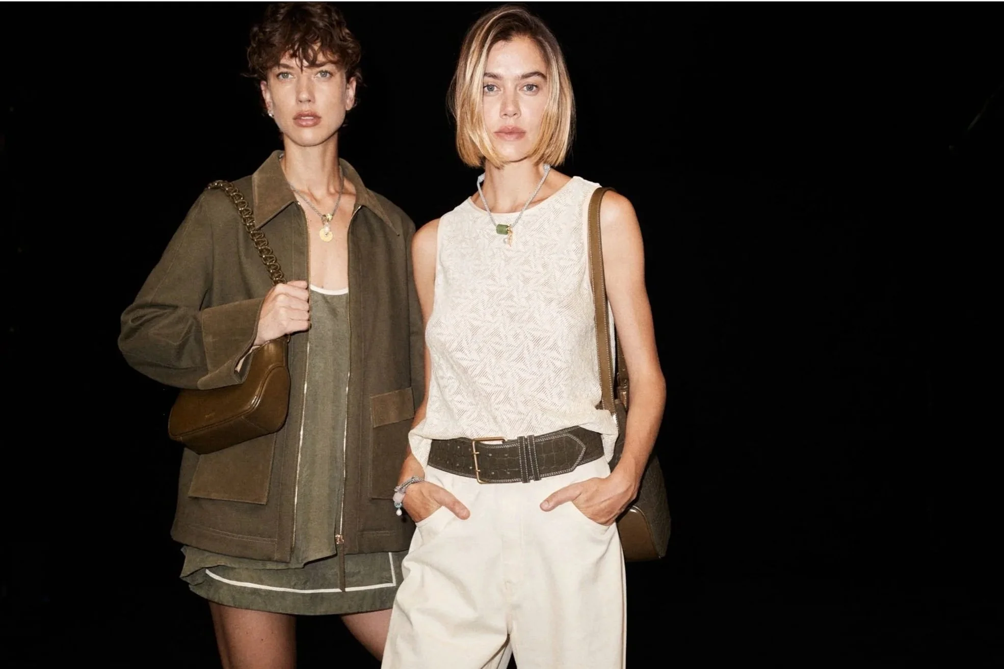 Two women standing against a black background, one wearing a dark olive green jacket and skirt with gold chain purse, the other in a sleeveless beige top and beige pants with a wide belt.
