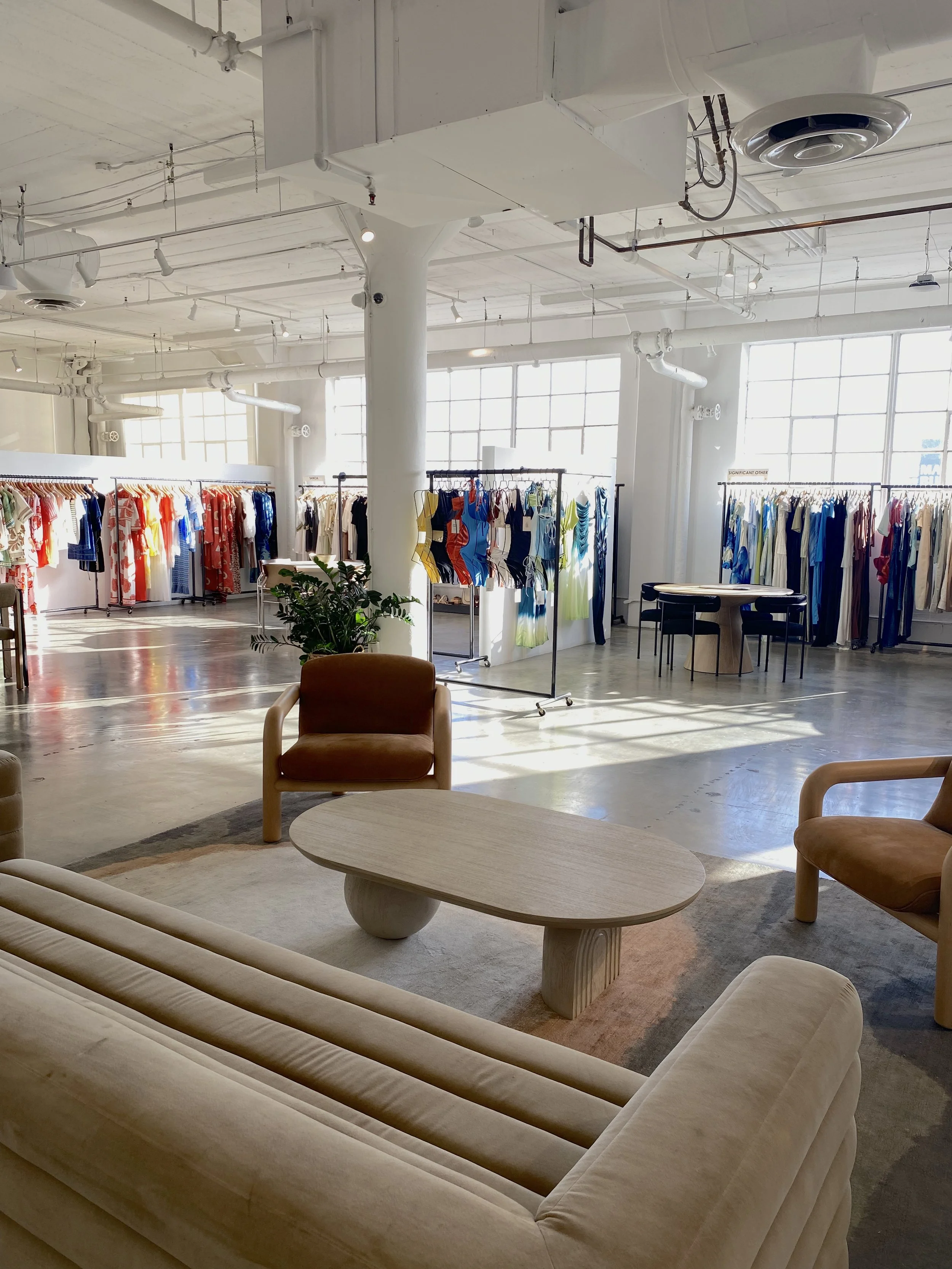 Modern, spacious clothing store with high ceilings, large windows, racks of colorful clothing, and a seating area with beige sofas and a low coffee table.