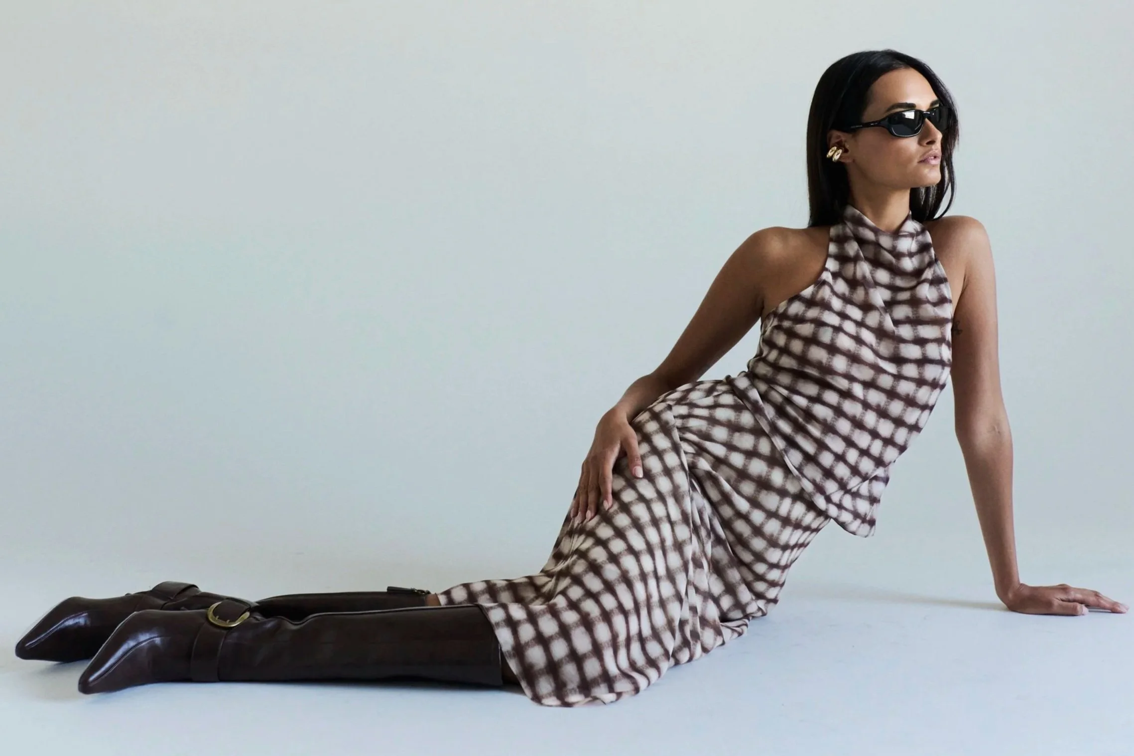 A woman with dark hair, sunglasses, and gold earrings posing on the floor in a checkered dress and black boots, with one hand on the ground and one knee bent, against a plain light-colored background.