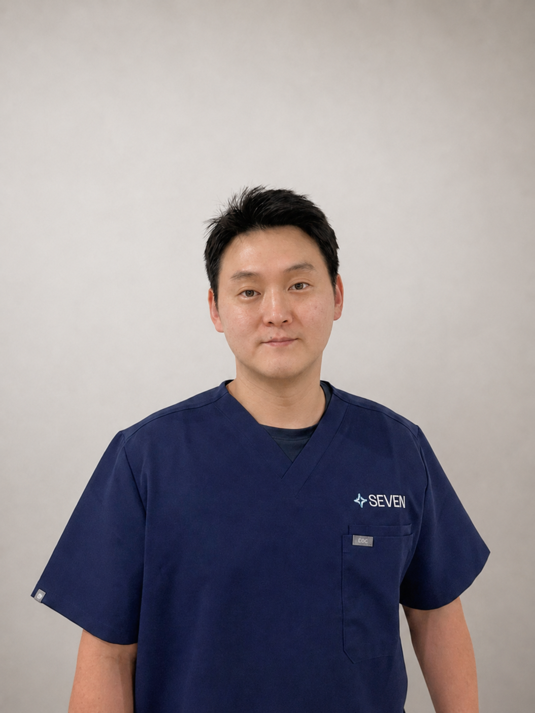 A man in navy blue scrubs with 'SEVEN' embroidered on the pocket, standing against a light gray background.