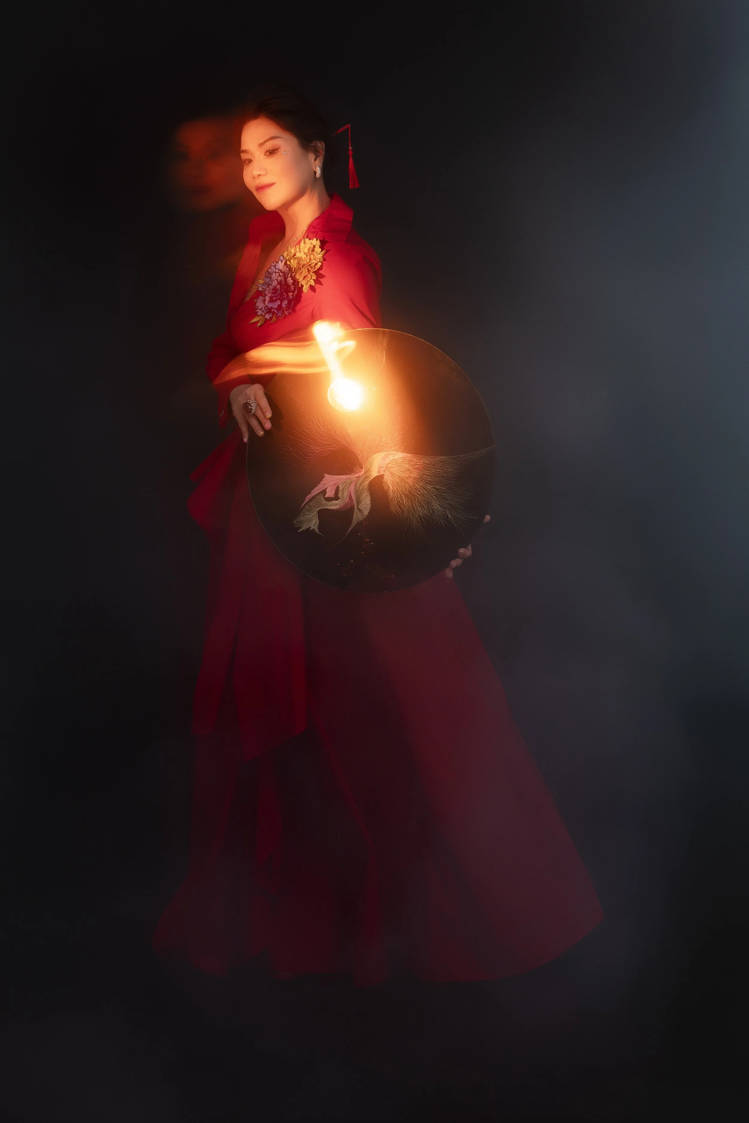 A woman in a long red dress holding a lamp with a glowing light, standing against a dark background. She is wearing earrings and a floral design on her dress.