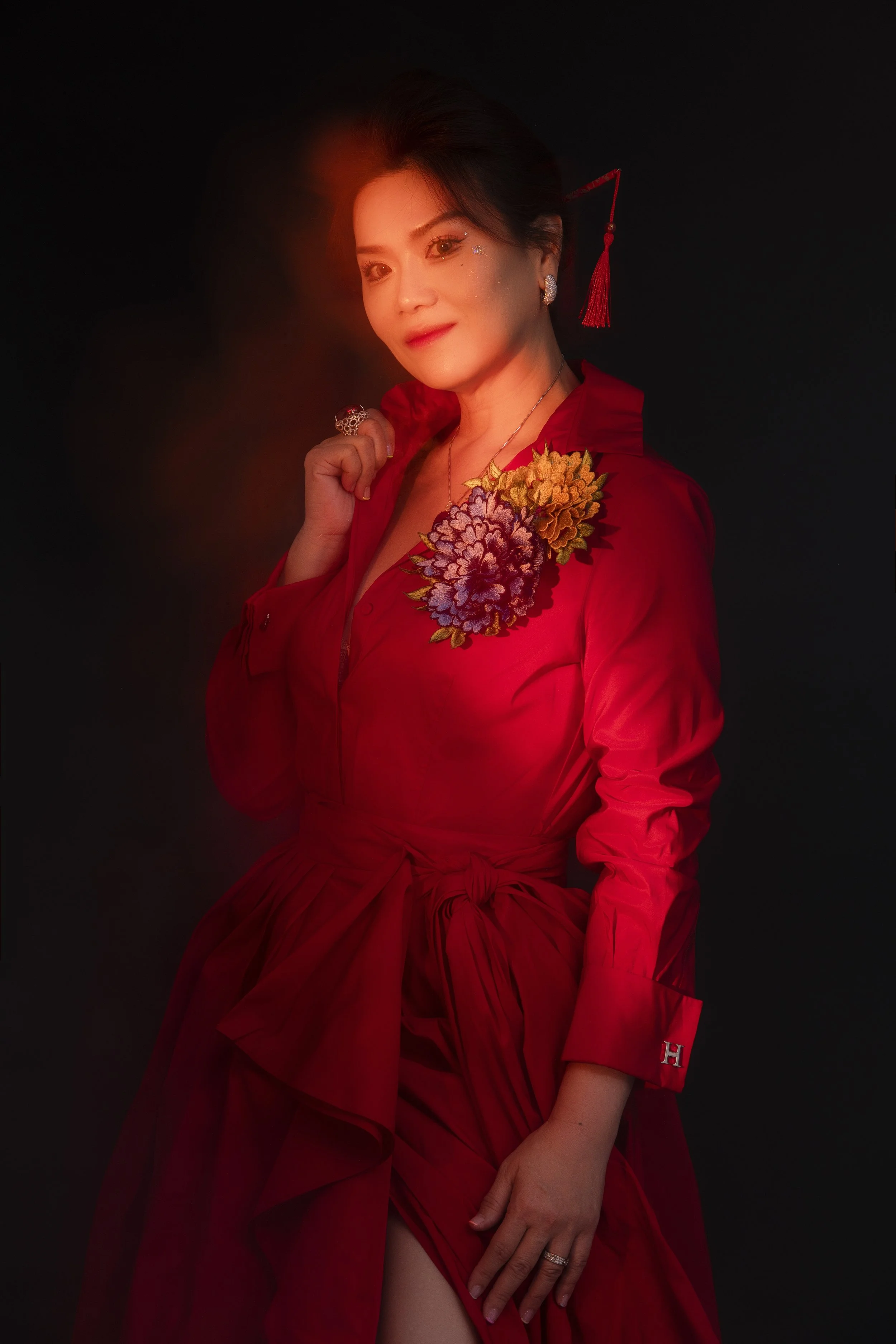 A woman dressed in a red outfit with floral embellishments, jewelry, and a red tassel earring against a dark background.