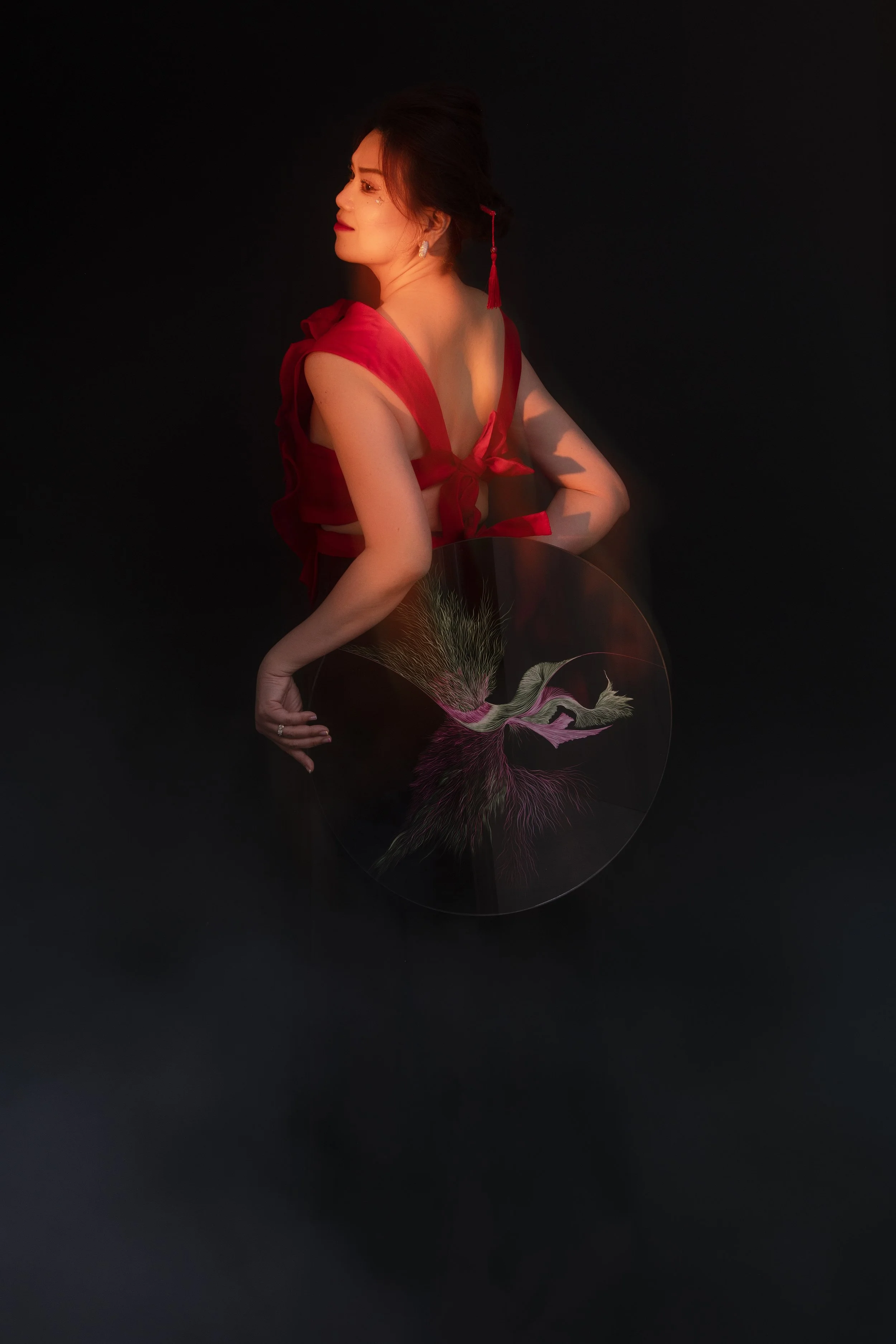 A woman with dark hair styled in an updo, wearing a red dress with off-the-shoulder ruffled sleeves, a dangling red tassel earring, and holding a circular object with an intricate floral design, against a dark background.
