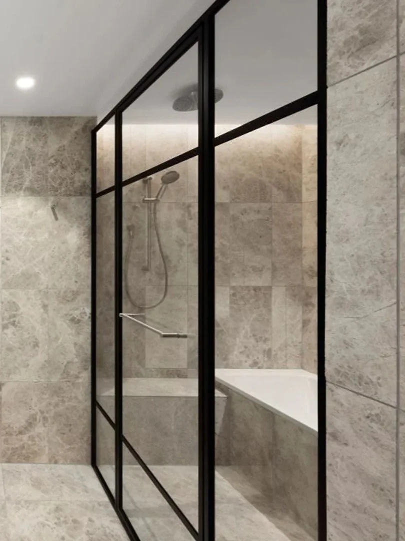 A modern shower enclosed with black-framed glass panels in a beige tiled bathroom, with a built-in bathtub inside.