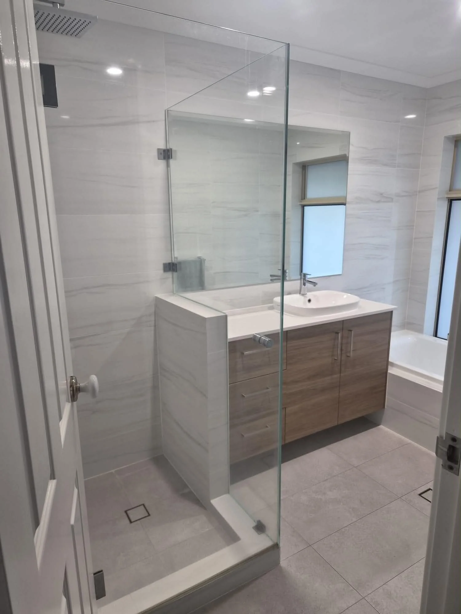 Modern bathroom with glass shower enclosure, white and wood vanity, and frosted windows.