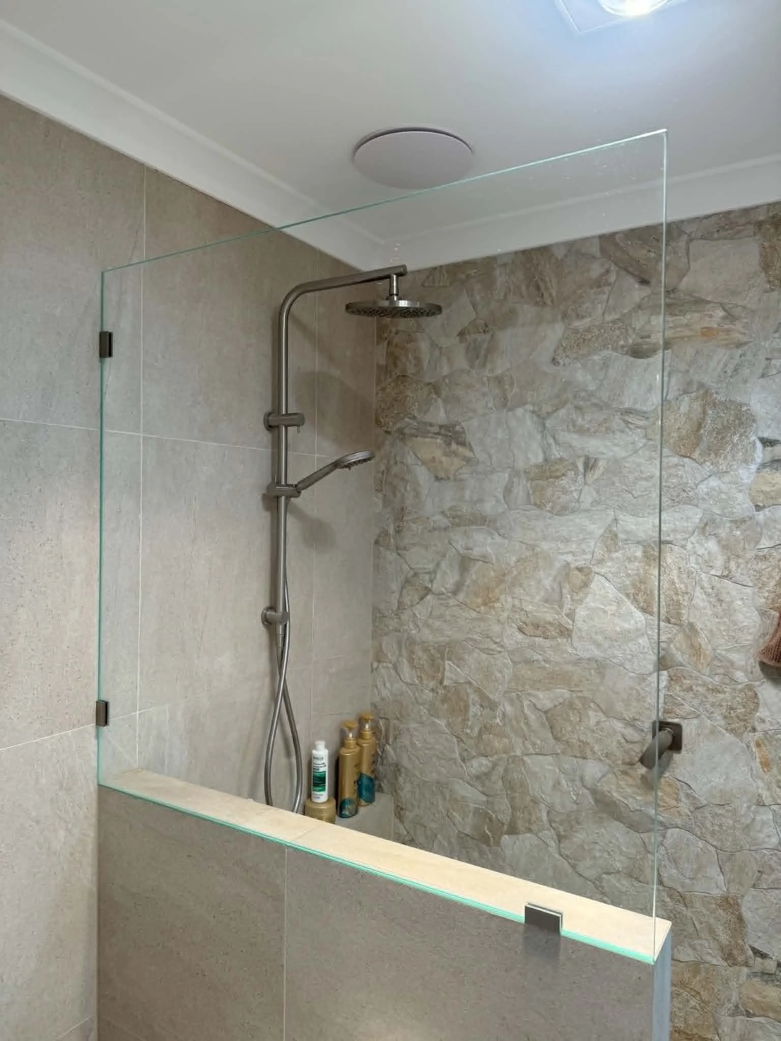Modern shower with glass partition, metal showerhead, beige and stone-tile walls, bottles of shampoo and soap on a ledge inside the shower.