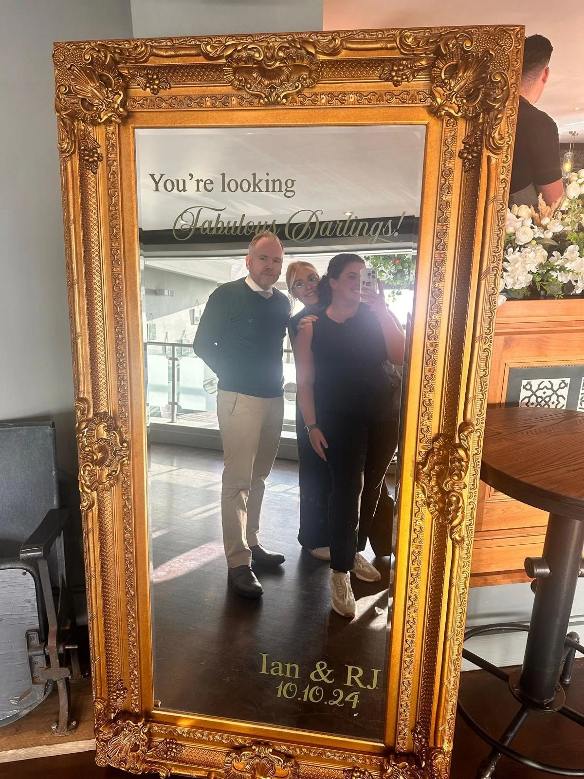 A large ornate gold-framed mirror with a message at the top reading, "You're looking Fabulous Darlings!" reflecting three people taking a selfie. The bottom of the mirror has the names Ian and RJ and the date 10.10.24. The scene appears to be in a we