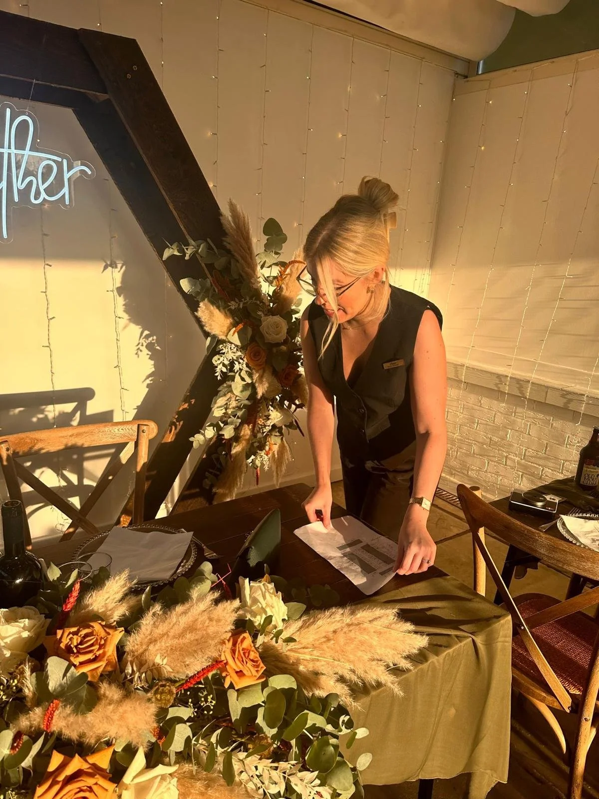 A woman wearing glasses and a black sleeveless top stands at a table, arranging papers. The table is decorated with a floral arrangement of roses and pampas grass, and there are wine bottles, glasses, and tableware nearby. Behind her, a decorative fl