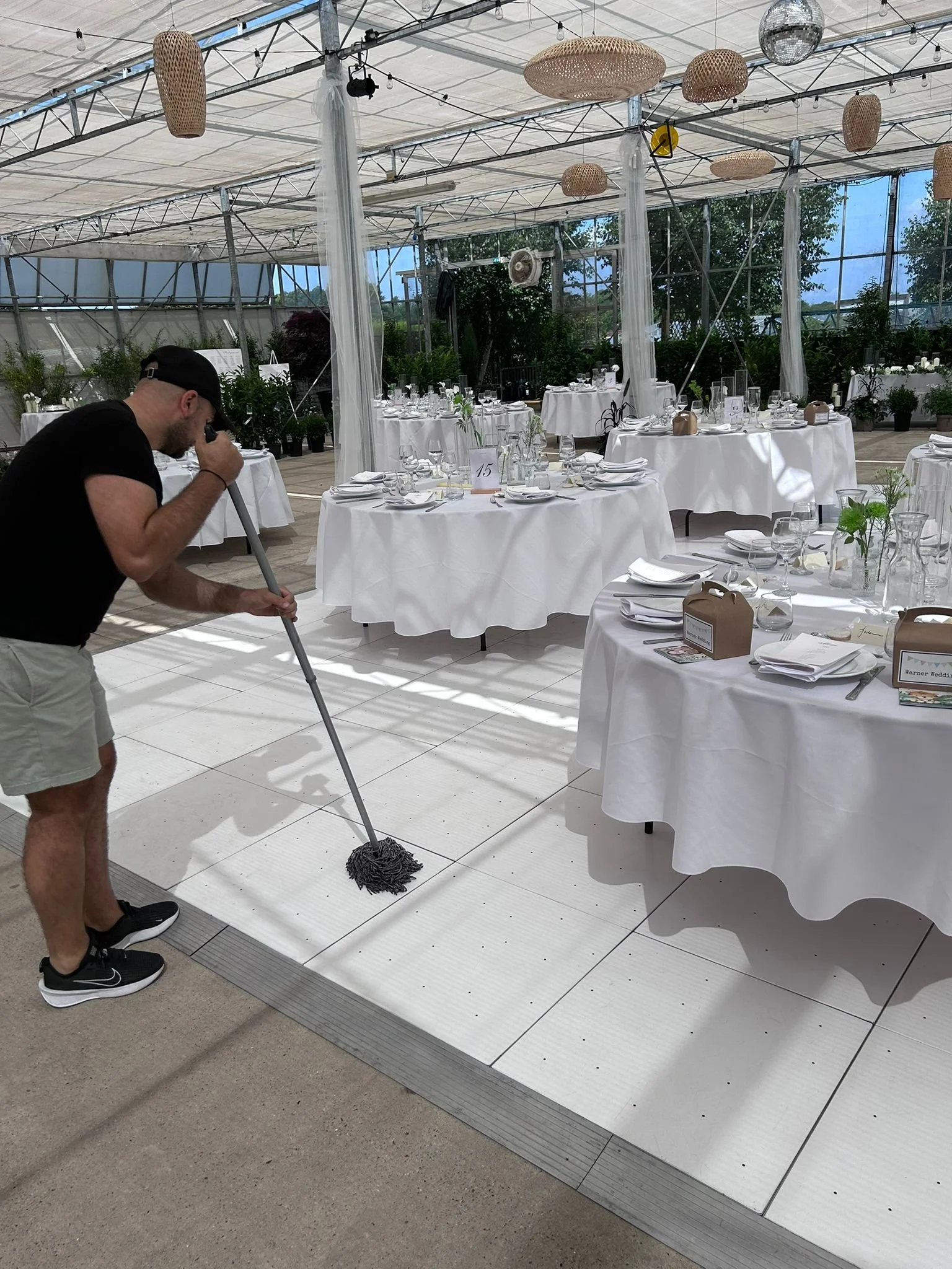 A man in black t-shirt, beige shorts, and black sneakers mops a white tile floor in a large event space with round tables draped in white tablecloths, set with plates, silverware, glasses, and small vases with greenery. The space is enclosed with gla
