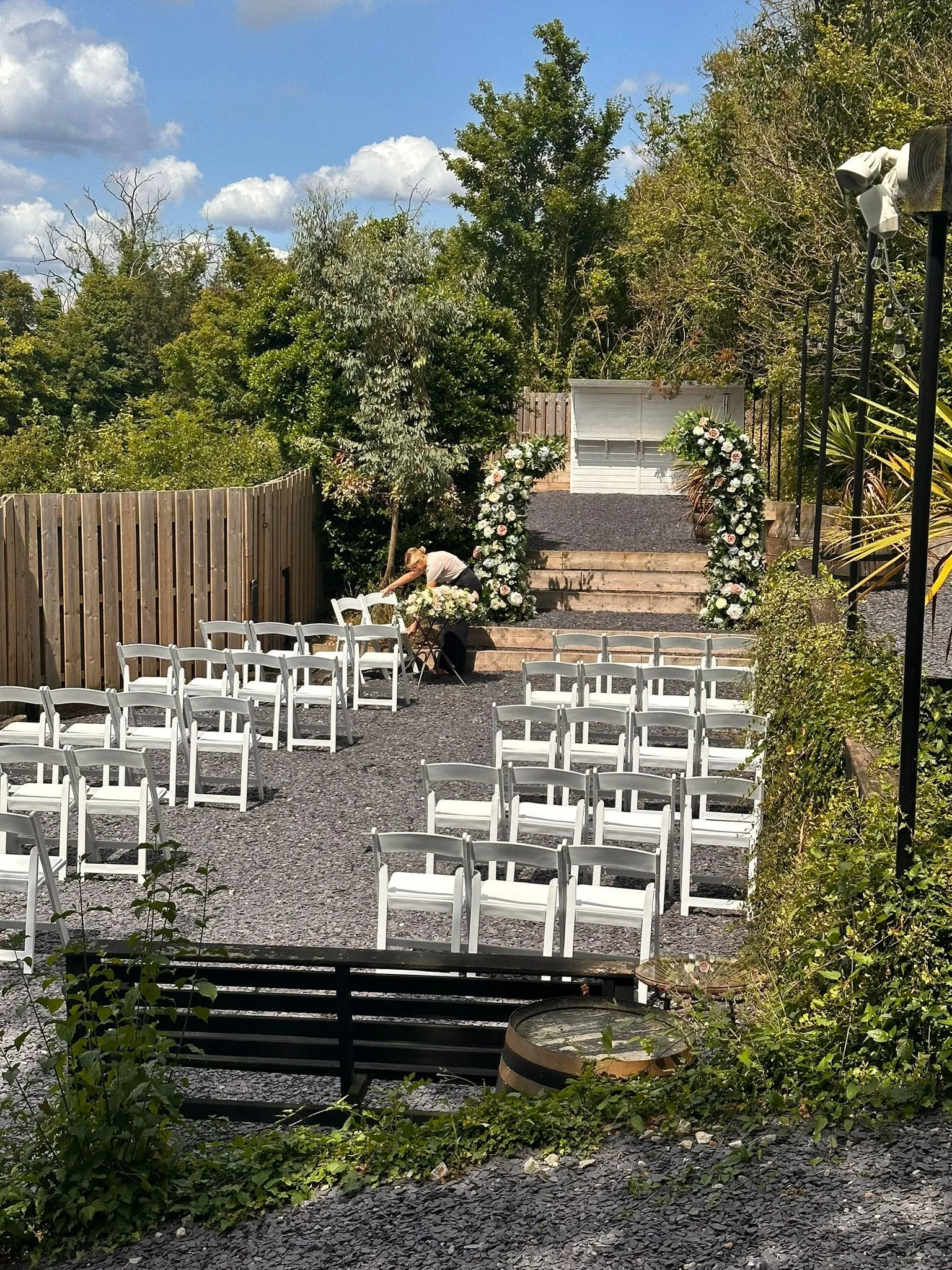 Outdoor wedding setup with white chairs arranged on a gravel ground, floral arch, and a backdrop of trees and blue sky.
