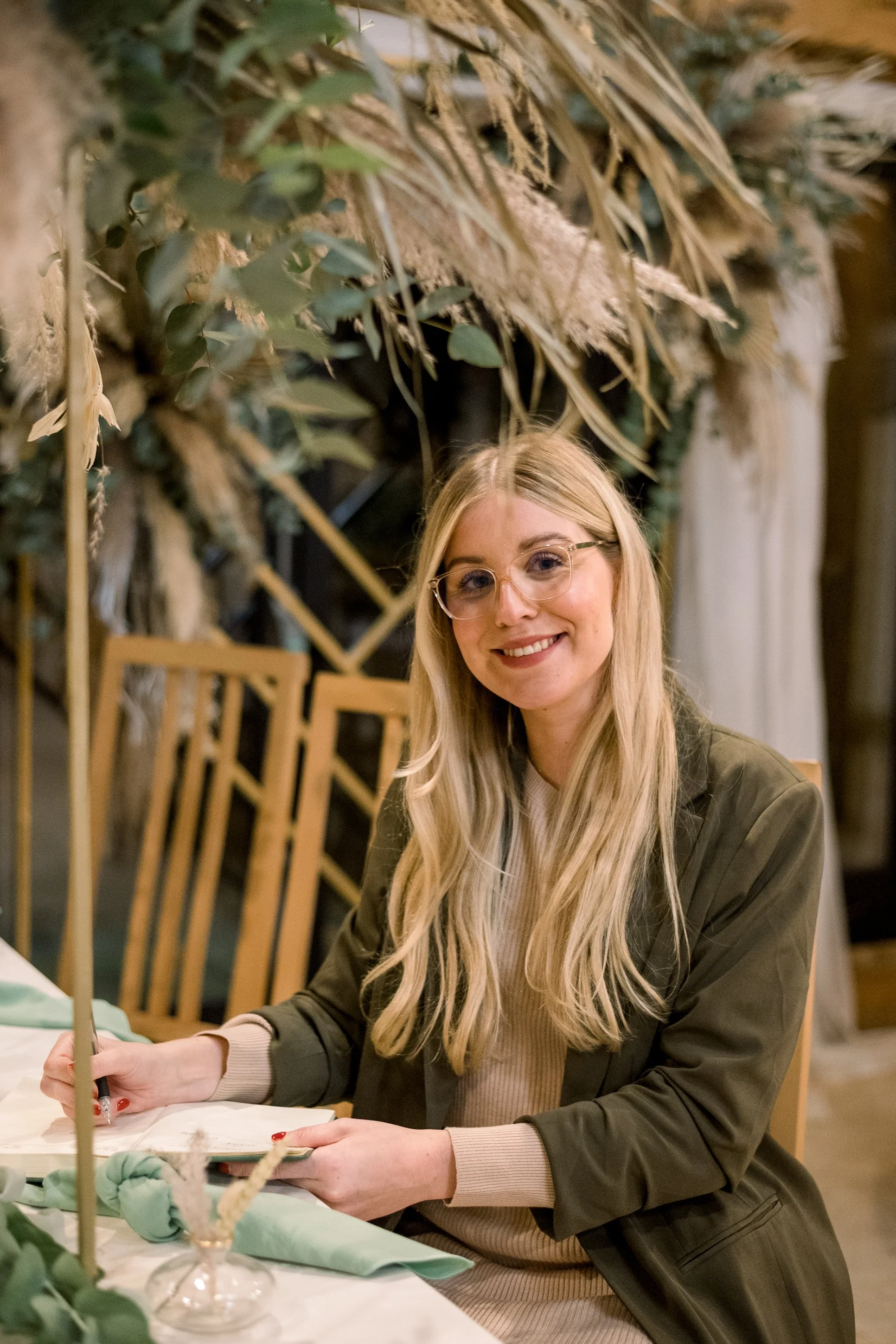 A smiling young woman with long blonde hair, wearing glasses and a dark jacket, sitting at a table with writing materials, in a decorated indoor setting.