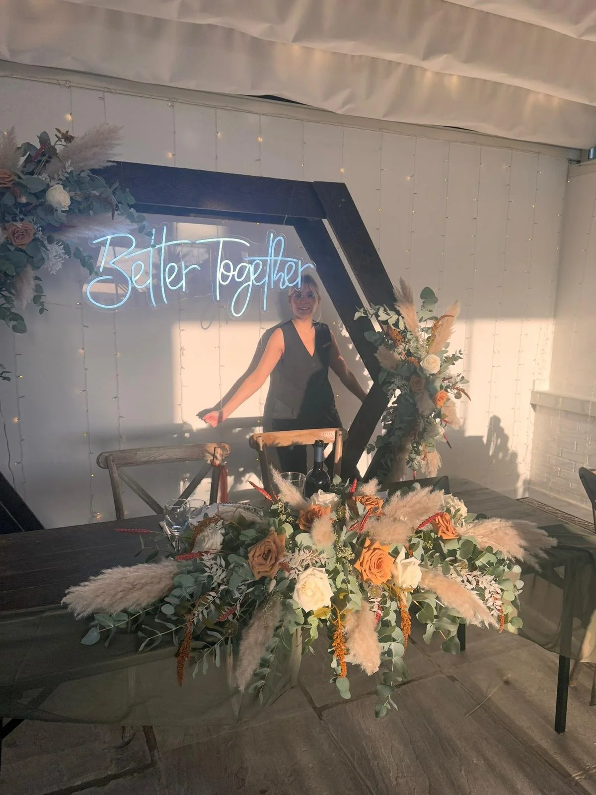 A woman smiling inside an event space with a neon sign that reads "Better Together." The space is decorated with floral arrangements and fairy lights, and there is a table with a wine bottle and glasses.