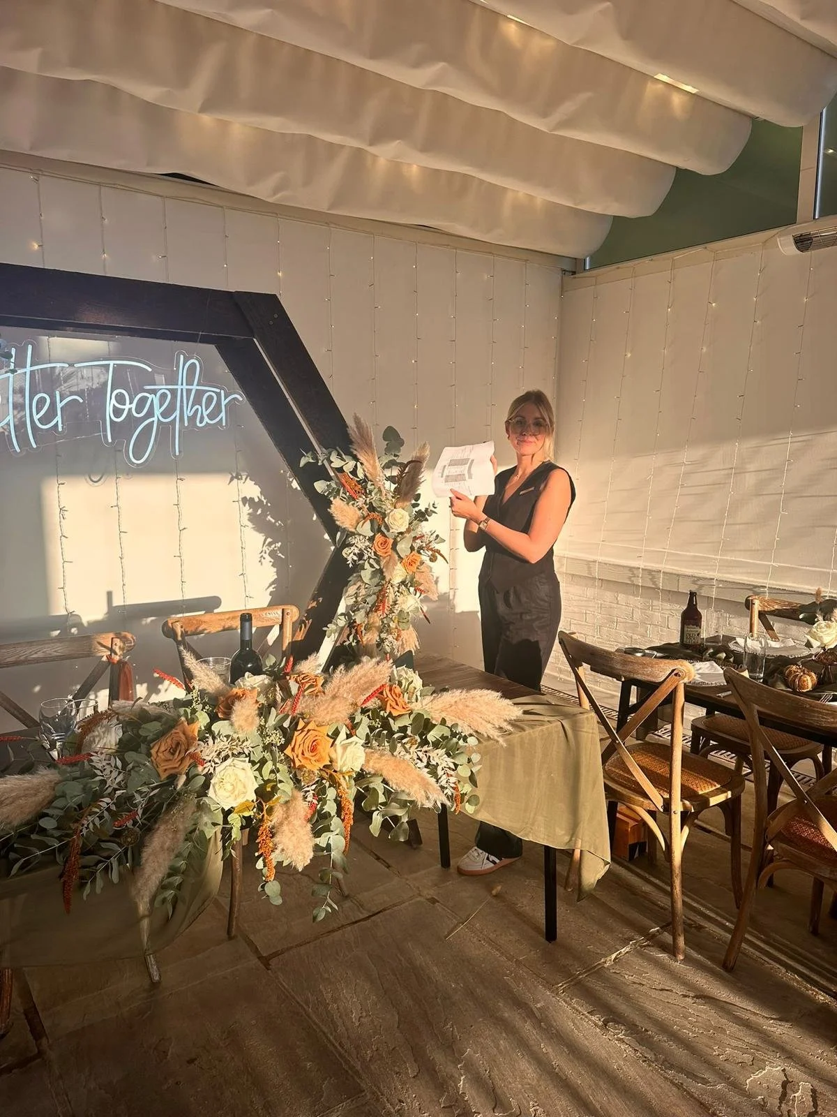 Woman standing at a decorated table with floral arrangements, holding a microphone or papers, in a well-lit room with white walls and a neon sign that says 'Better Together'.