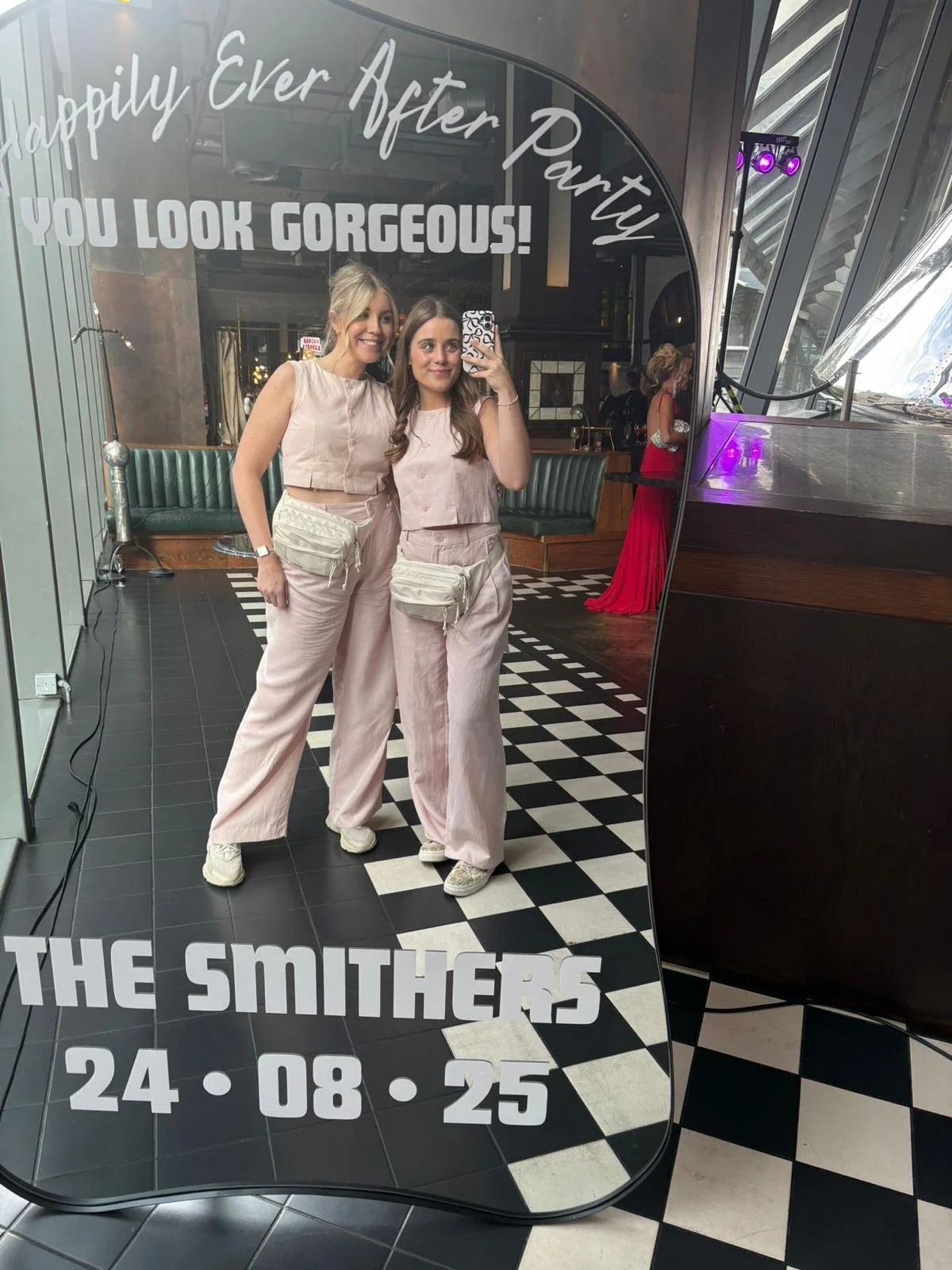 Two young women taking a selfie in front of a mirror inside a restaurant or bar. They are wearing matching light pink outfits and fanny packs. Behind them, there is another woman in a red dress. The mirror has text that reads, "Happily Ever After Par