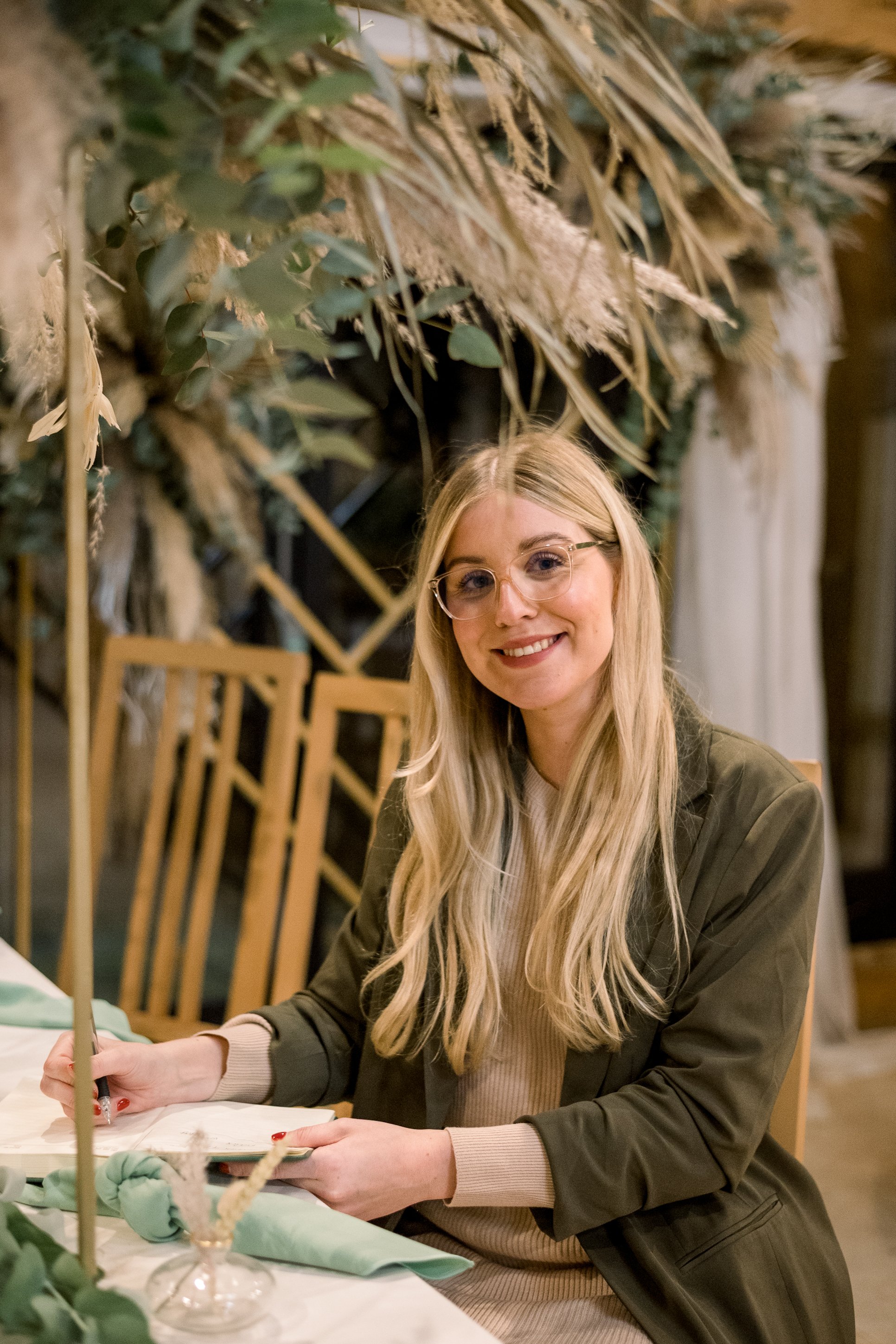 A young woman with long blonde hair and glasses sitting at a table, smiling, in a setting with a decorated background, writing in a notebook.