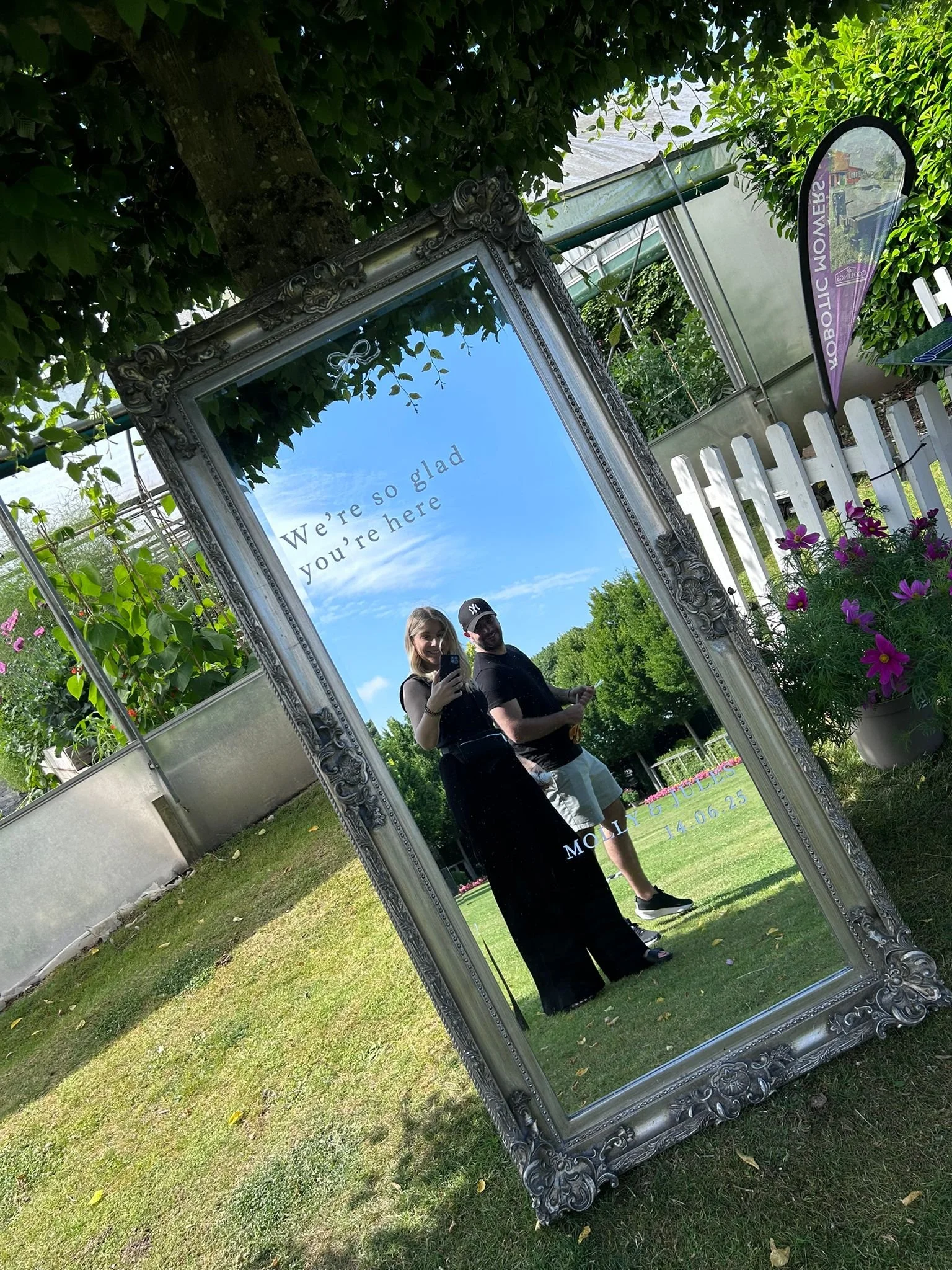 A large ornate mirror leaning against a tree in a garden. The mirror shows a reflection of a woman and a man taking a photo together. The woman has blonde hair and is wearing a black dress; the man is wearing a black shirt with a white logo cap. Ther