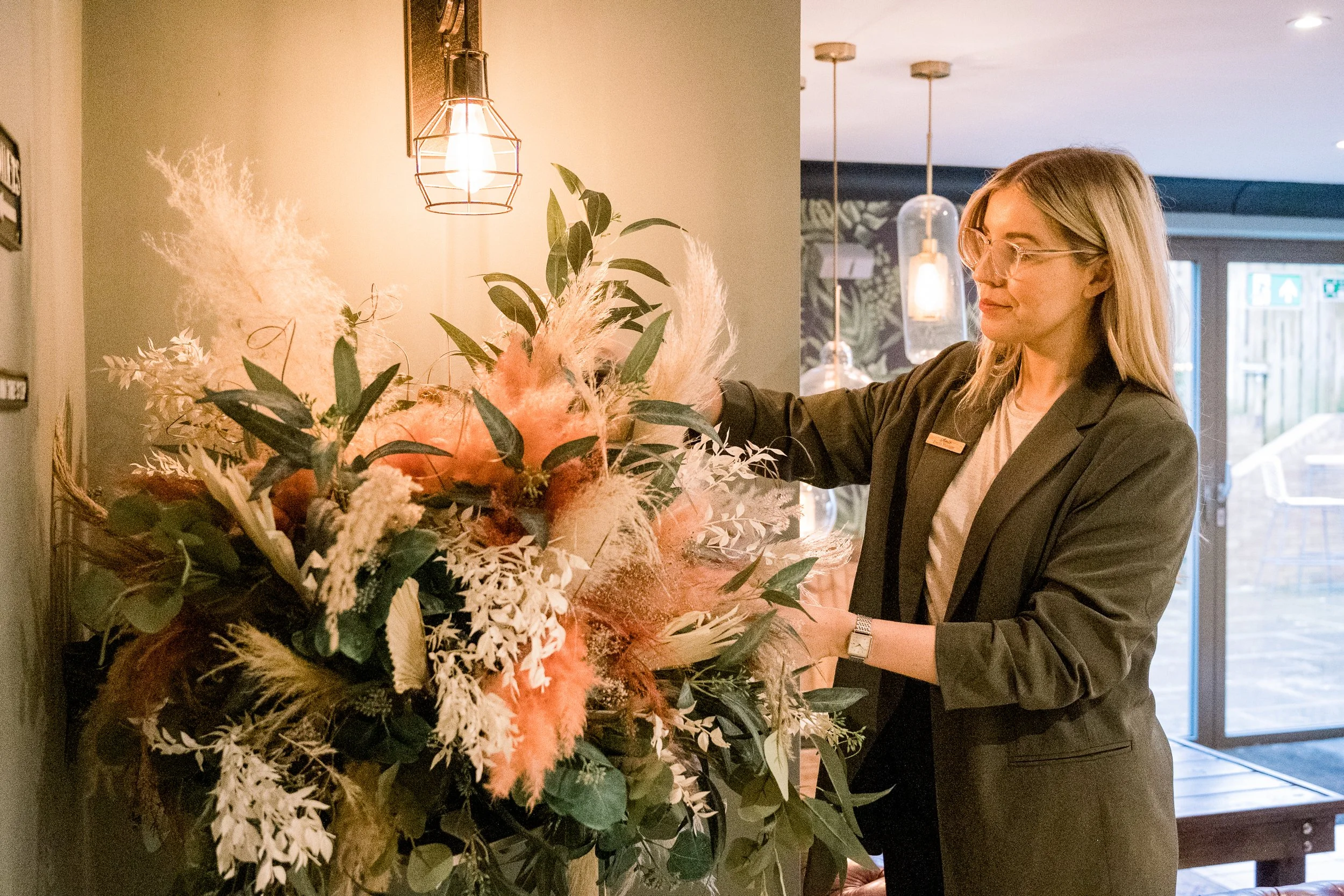 A woman wearing glasses and a blazer arranges a large bouquet of flowers and greenery inside a well-lit room with hanging pendant lights and a sliding glass door in the background.