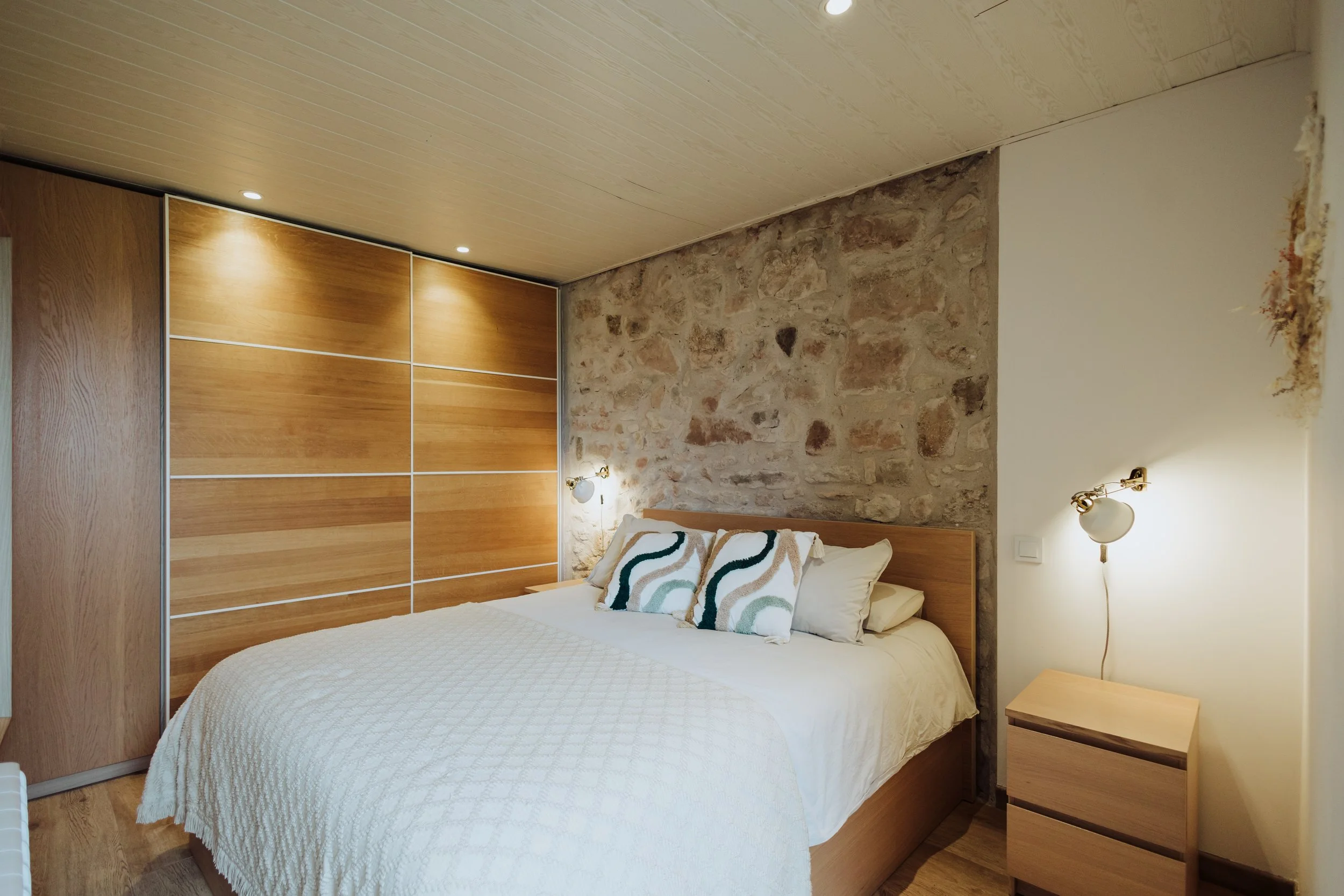 A bedroom with a bed that has a white quilt and decorative pillows, a wooden nightstand, wall-mounted lamp, exposed stone wall, wooden closet doors, and a beige ceiling with recessed lighting.