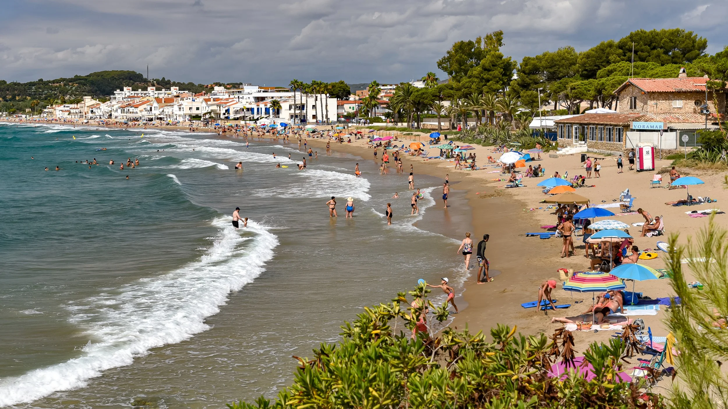 A lively beach scene with many people sunbathing, swimming, and walking along the sandy shore. Colorful umbrellas and beach towels are scattered across the sand, and there are buildings and trees in the background under a cloudy sky.