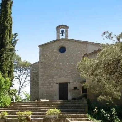 Small stone church with a bell tower, set on steps surrounded by trees and greenery under a clear blue sky.
