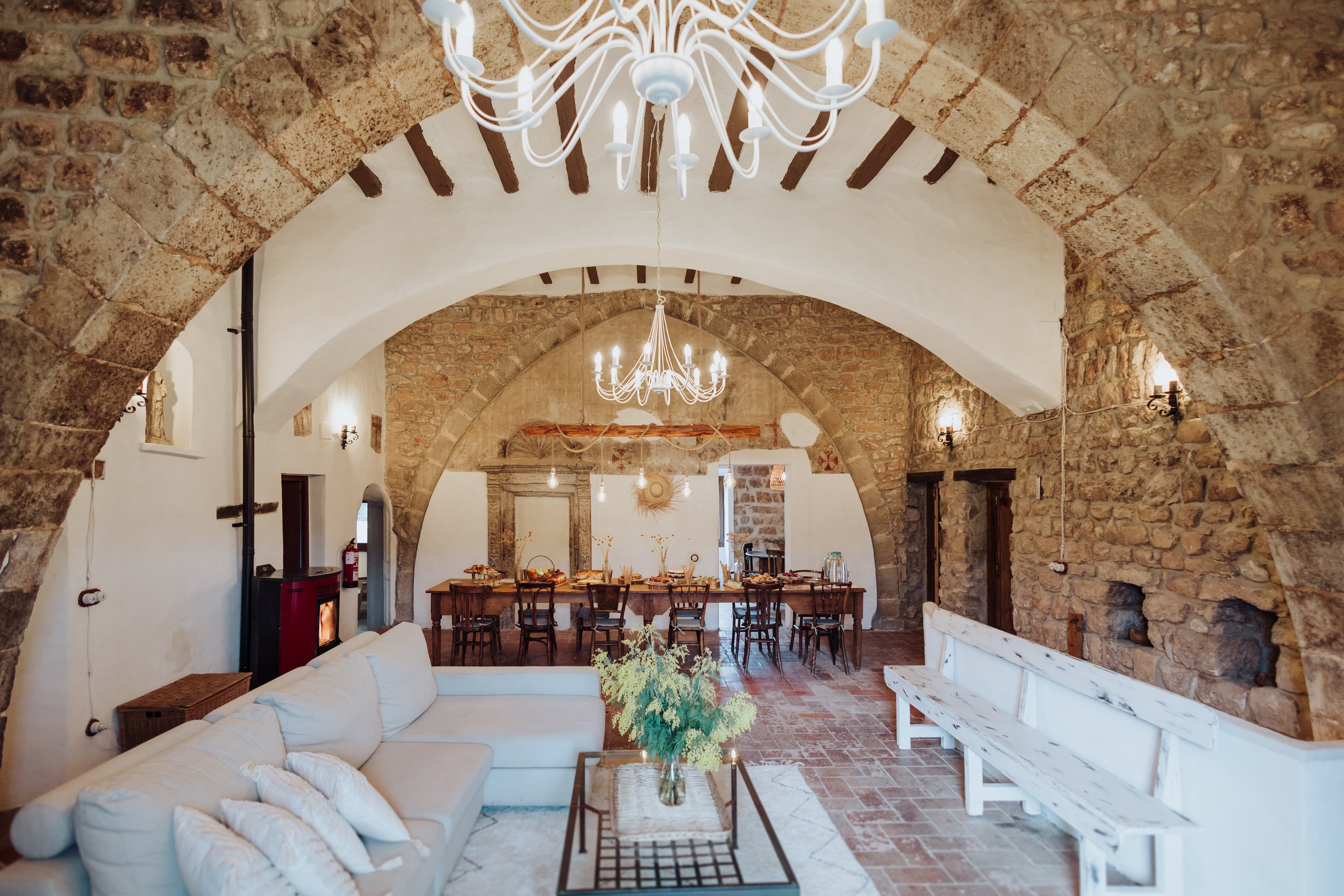 A cozy dining area in a rustic, stone-vaulted room with white walls and wooden beams. A white sofa with pillows is in the foreground, and a wooden dining table with chairs is in the background. Elegant white chandeliers hang from the ceiling, illuminating the space.