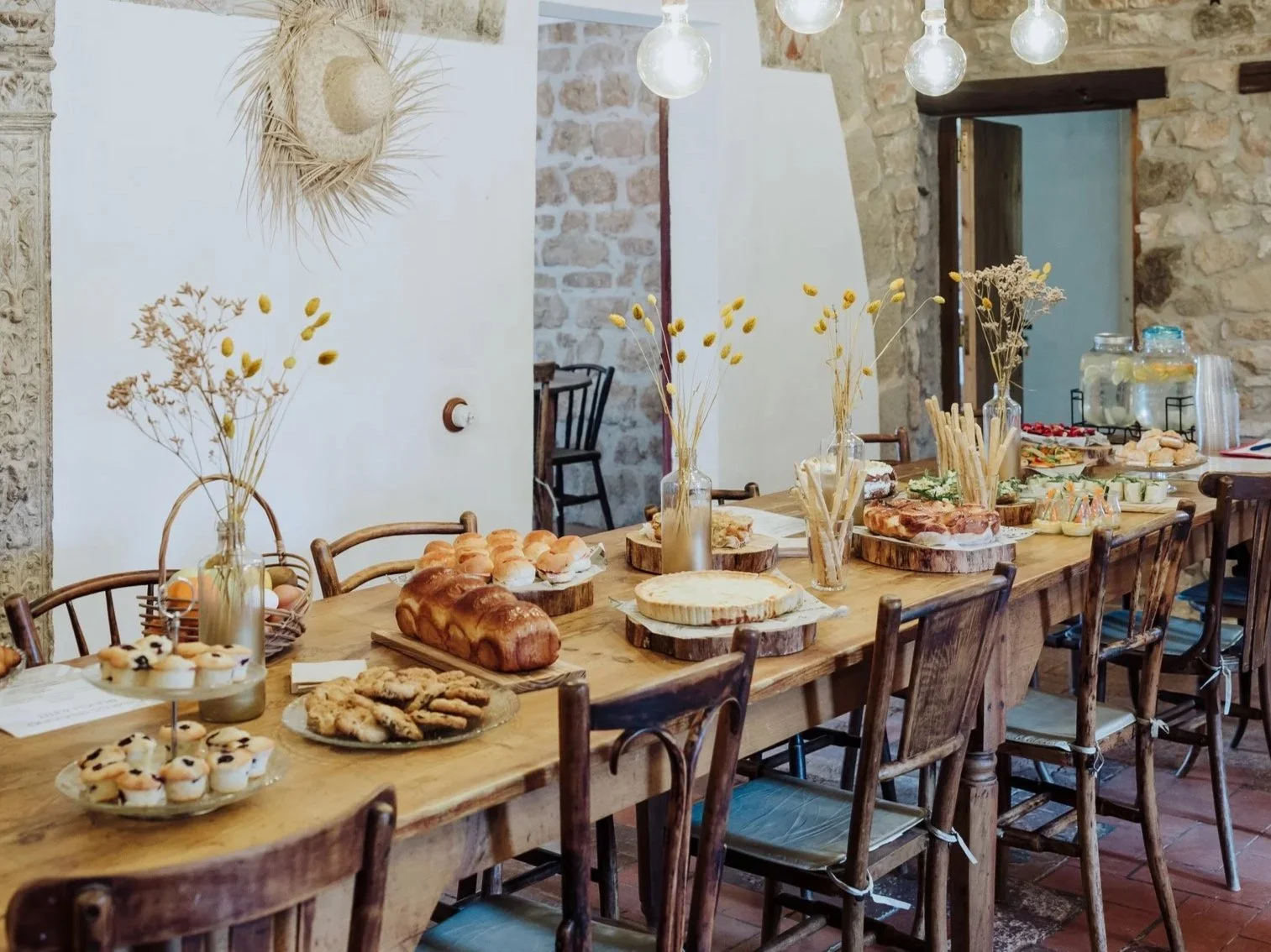 A rustic wooden table set for brunch with various baked goods including bread, cookies, and pastries, decorated with vases containing dried flowers, in a room with stone walls and hanging light bulbs.
