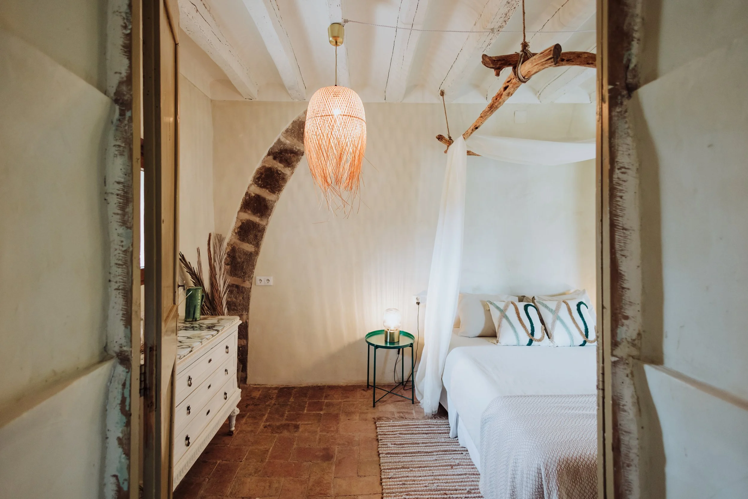 A cozy bedroom with a rustic style, featuring a white bed with patterned pillows, a small green side table with a lamp, a vintage white dresser, a hanging woven pendant light, and exposed wooden beams on the ceiling.