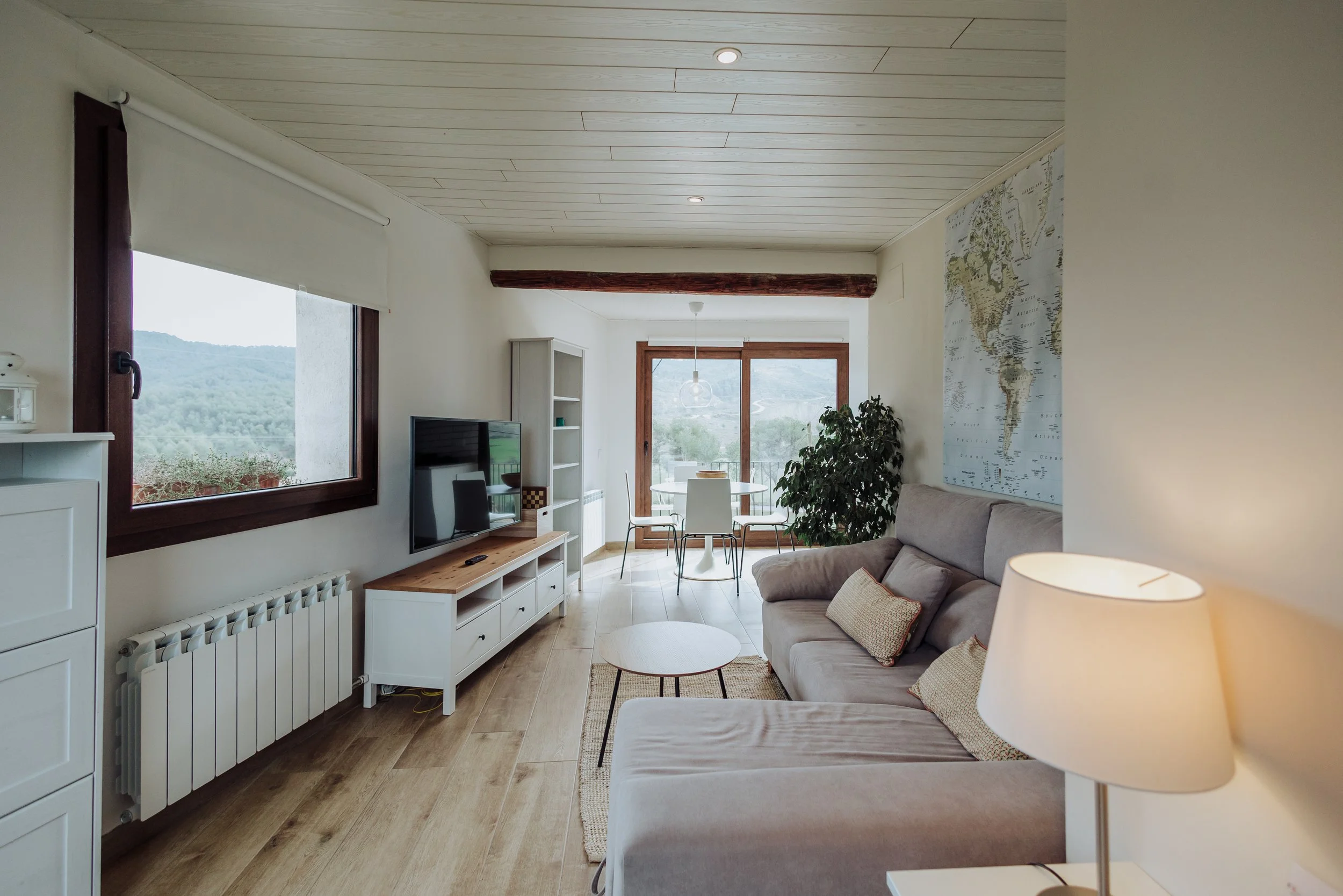 Living room with large window overlooking a wooded landscape, beige sofa with throw pillows, white entertainment center with TV, round glass dining table with white chairs, world map on wall, hardwood flooring, wood beam on ceiling, rattan rug, potted plant near window, ceiling lights, and sliding glass door leading to balcony.