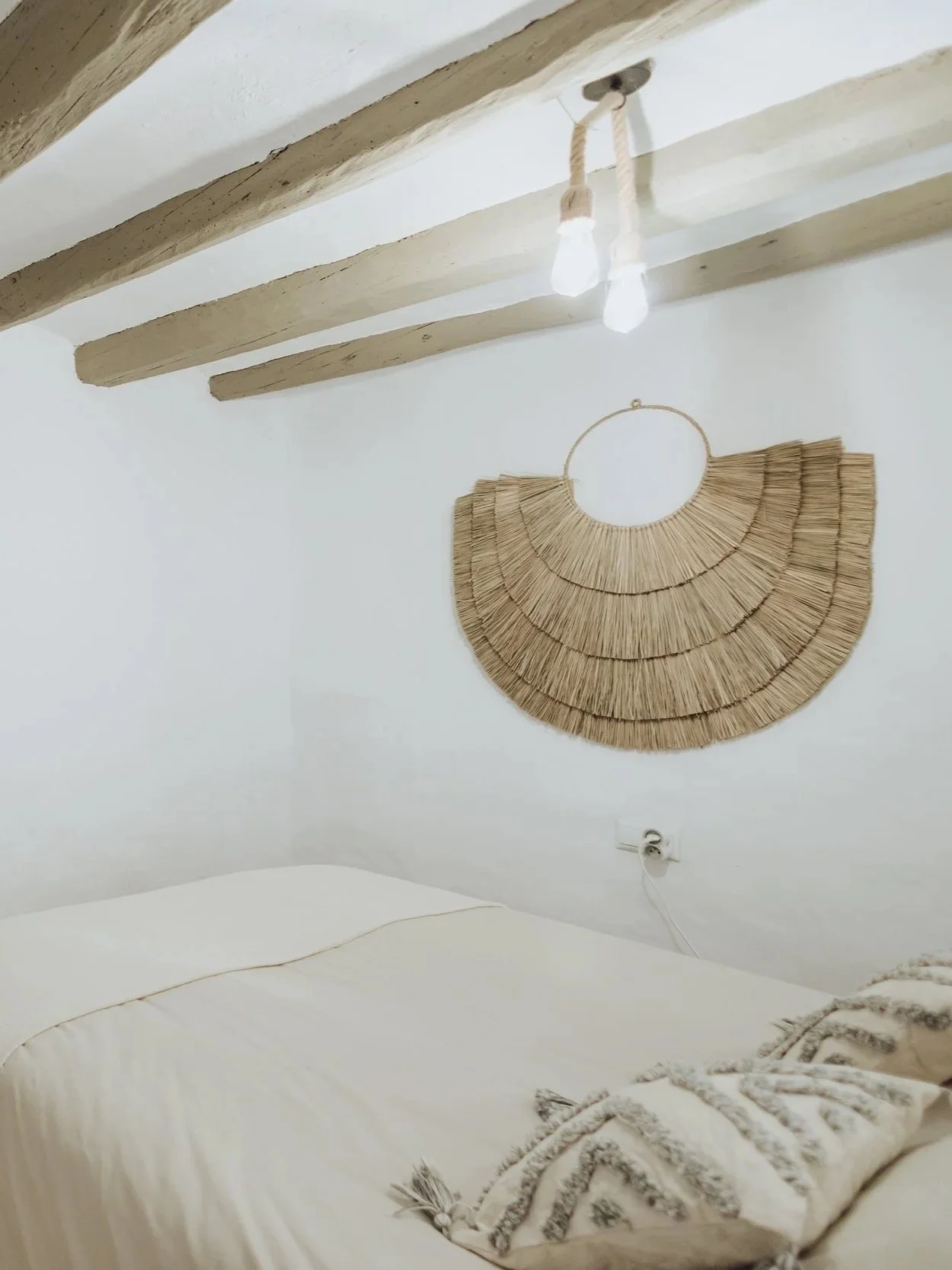 A cozy bedroom with white walls and wooden ceiling beams. A woven half-circle wall hanging decorates the wall above the bed, which has a beige blanket and a decorative pillow with tassels.