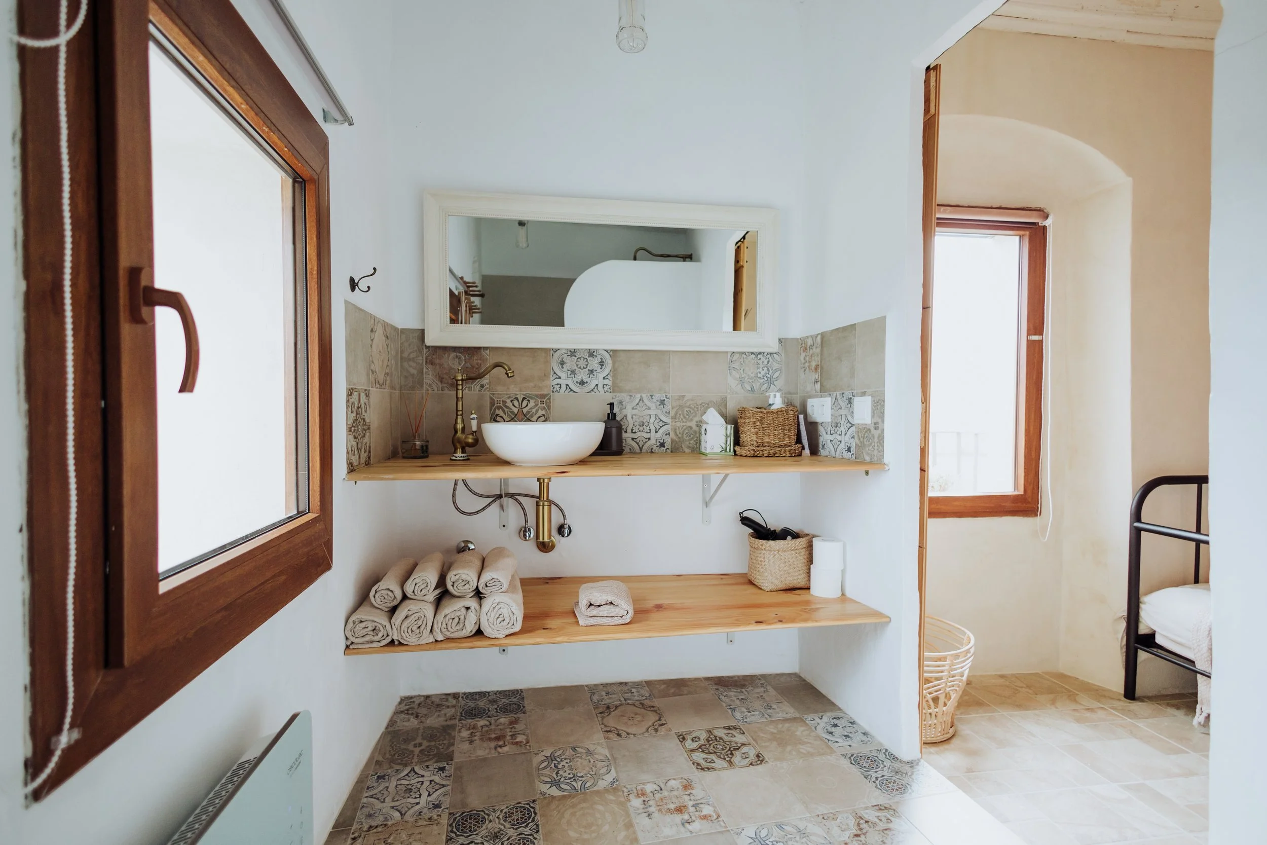 A bathroom with a wooden window, white walls, a large mirror, a vessel sink with a brass faucet, and a wooden shelf underneath holding rolled towels and other bathroom essentials. The floor is tiled with patterned tiles, and there is a wicker basket in the corner near a bed with black metal frame. A small window lets in natural light.