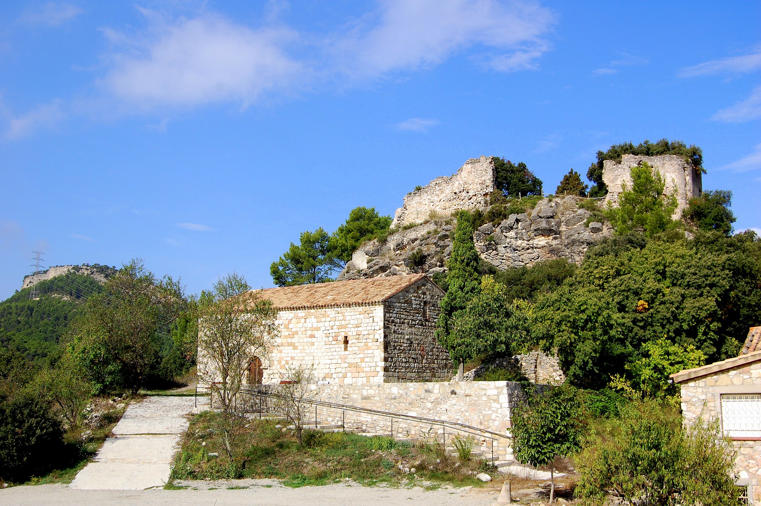 Ruins of an ancient stone castle on a hilltop surrounded by green trees under a bright blue sky with scattered clouds.