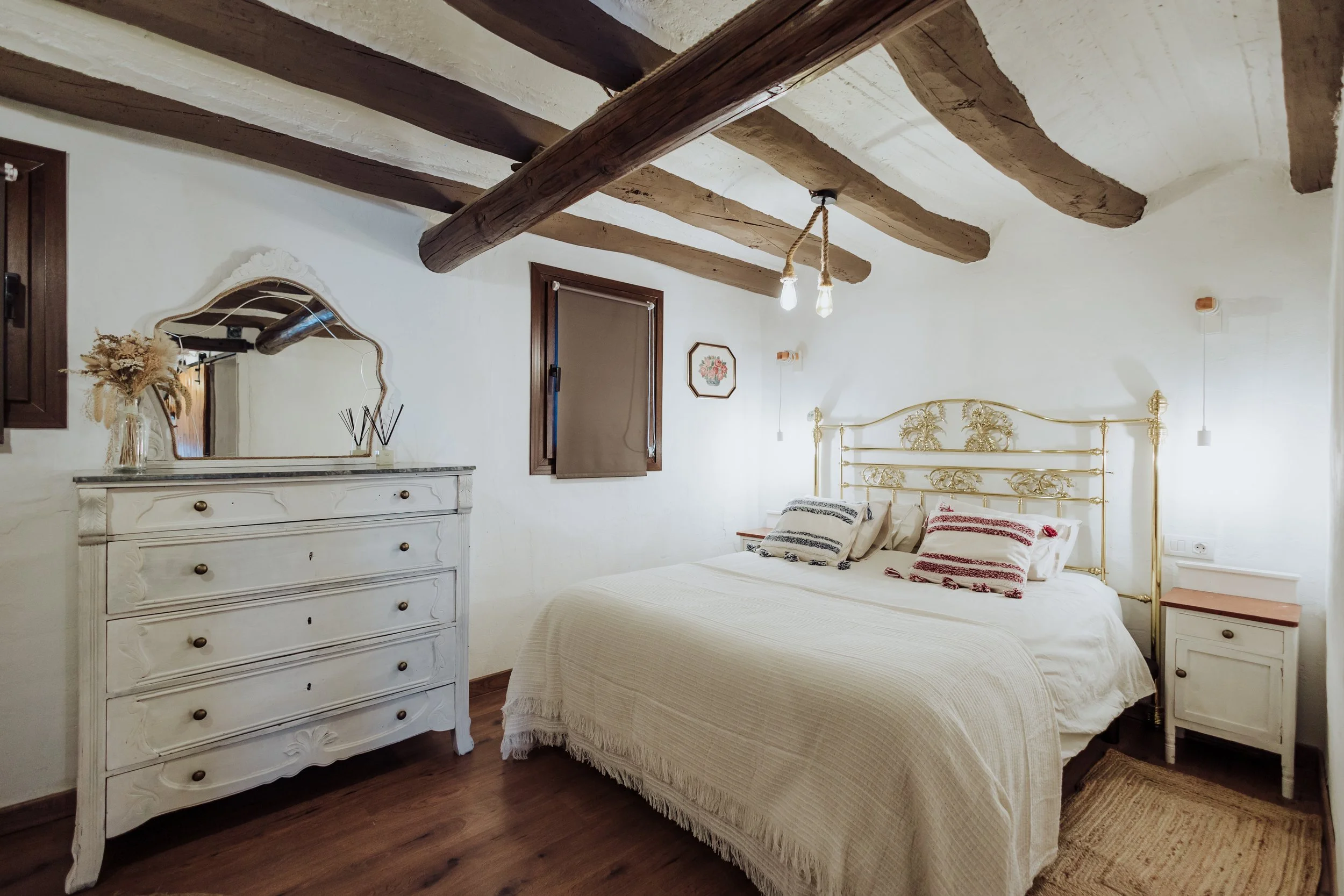 A cozy bedroom with a white vintage dresser, a decorative mirror, and a bed with a gold ornate headboard, white bedding, and striped pillows. The room features exposed wooden ceiling beams, wooden window frames, and hanging light bulbs, with dried flowers in a vase on the dresser.