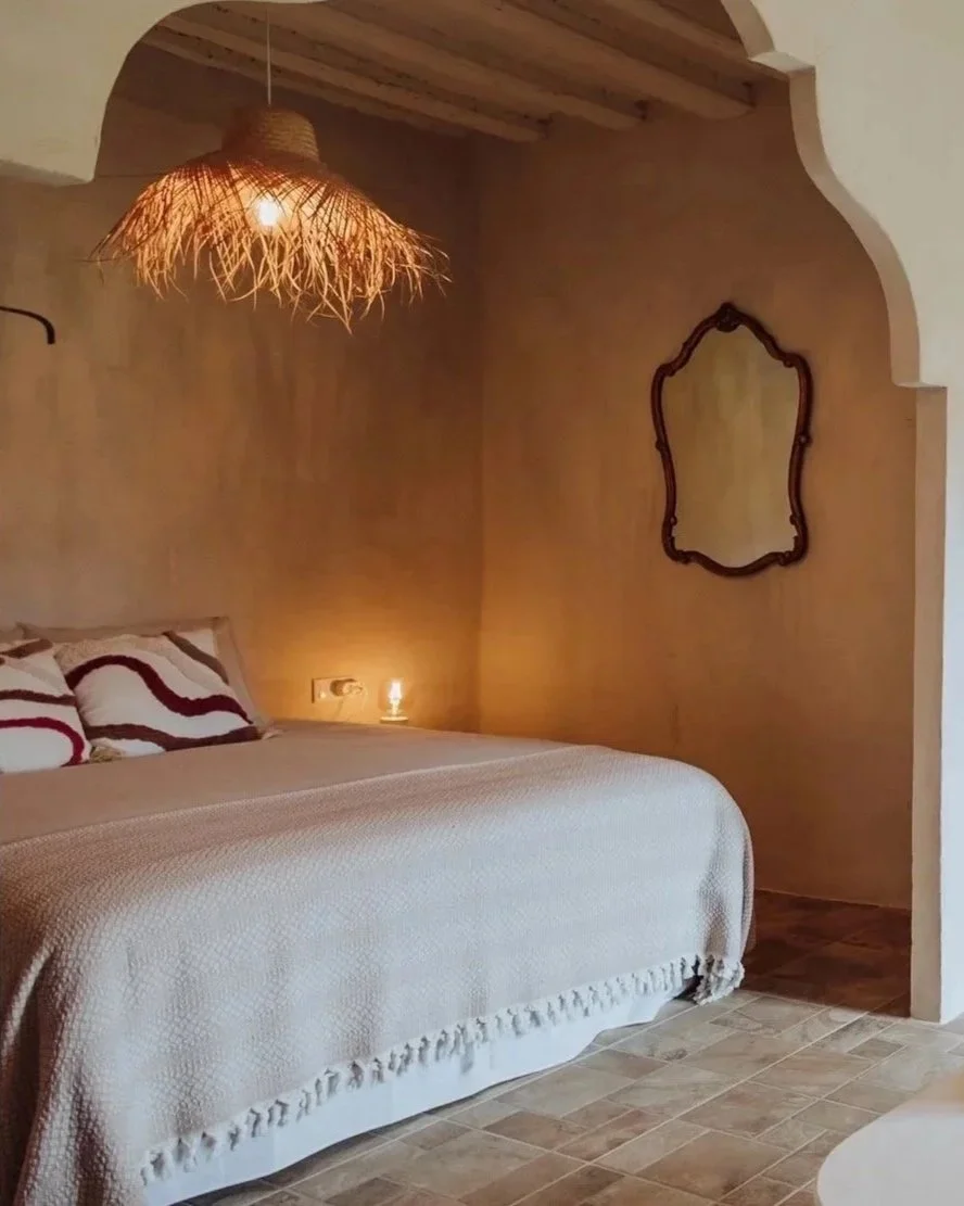 A cozy bedroom with a beige bedspread, decorative pillows with red and black wavy patterns, a woven hanging lamp, wall mirror with dark frame, and warm lighting.