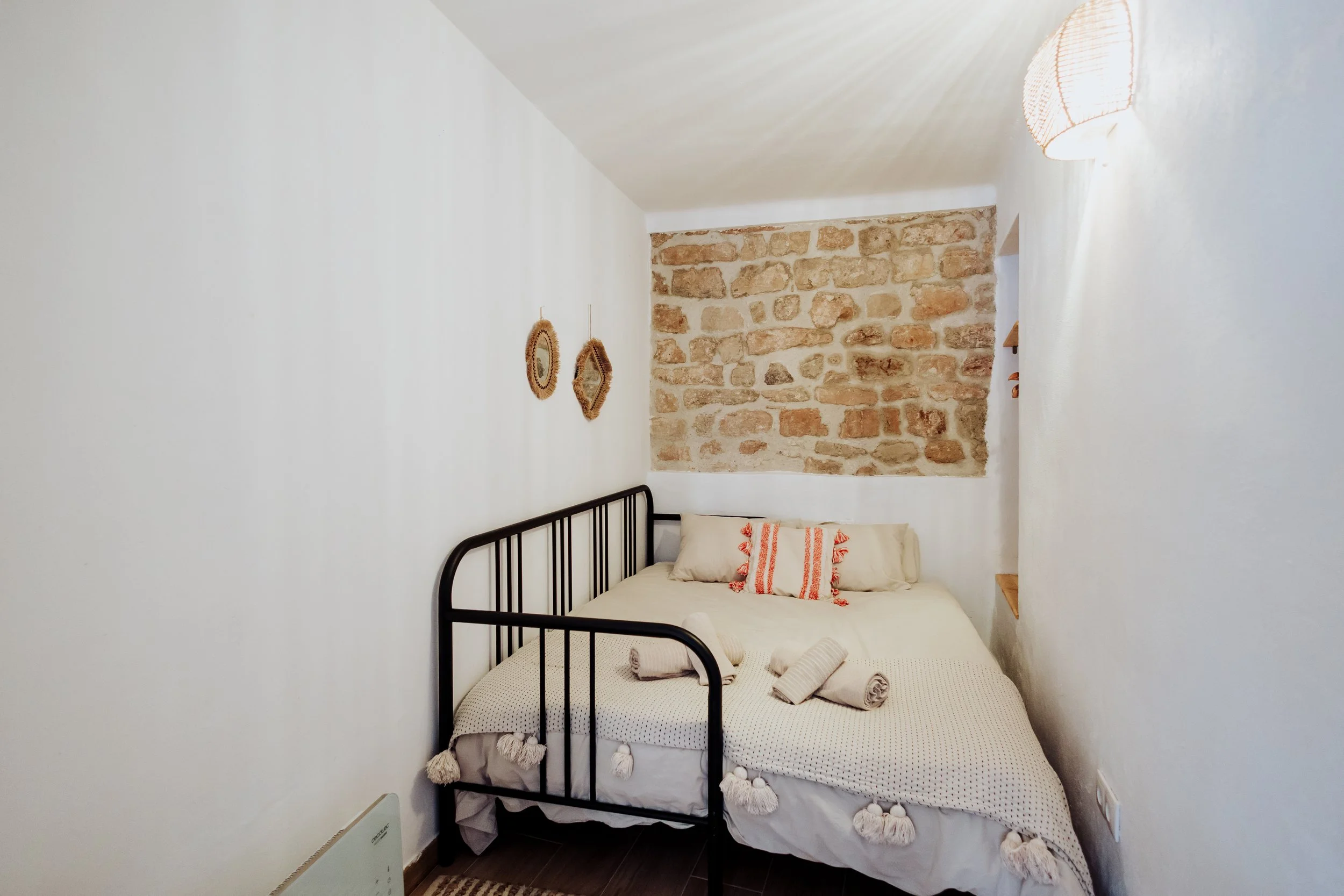 A bedroom with white walls, a stone accent wall, a black metal bed frame, a bed with white bedding and red-striped pillows, and a rattan wall decoration.
