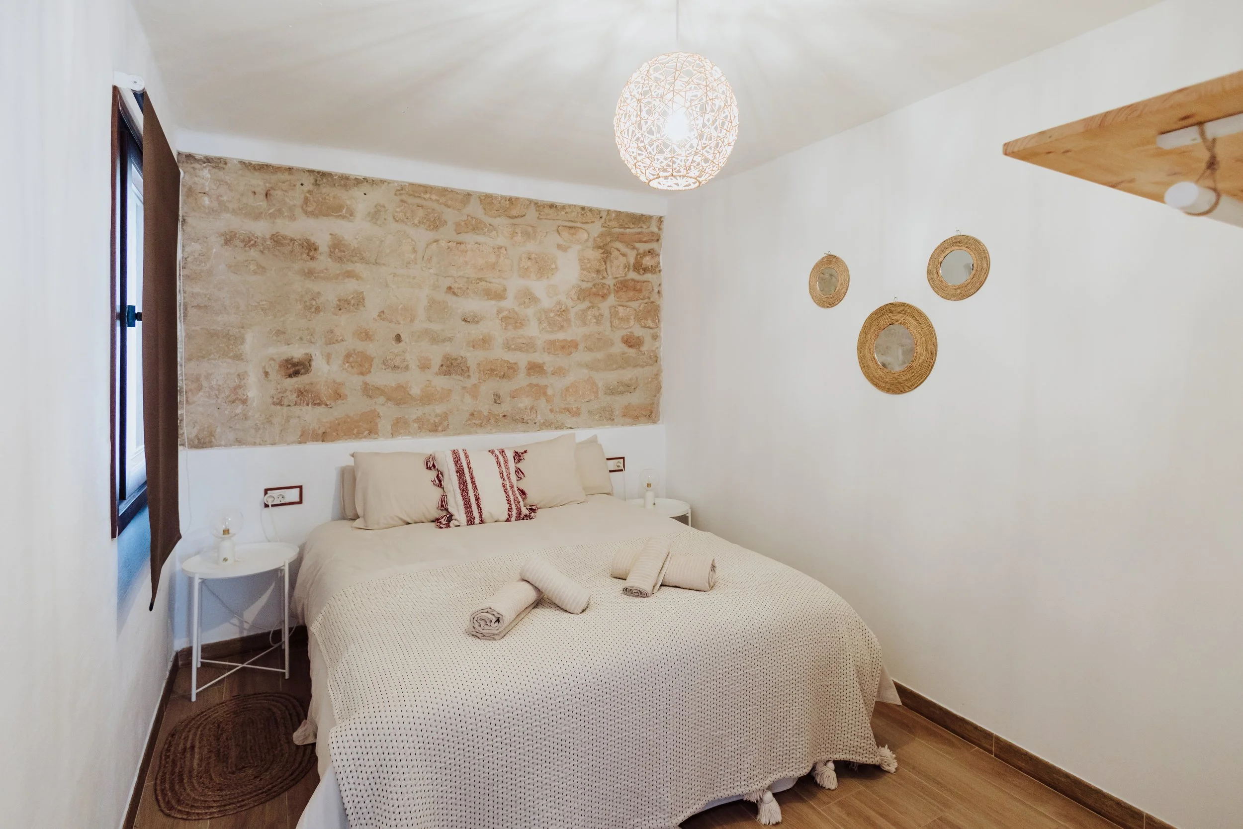 A cozy bedroom with a white bed, beige bedspread, and rolled towels on top, flanked by two bedside tables; a stone accent wall behind the bed; a woven light fixture hanging from the ceiling; two small round mirrors on the white wall; hardwood floors; a window with dark curtains on the left side.