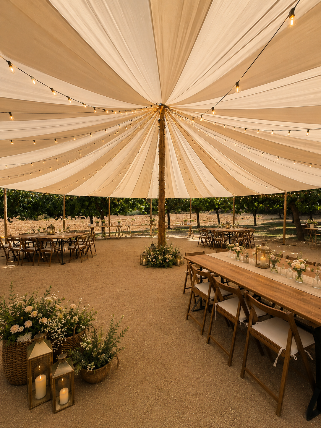 Outdoor event setup under a large beige fabric tent with string lights, wooden tables decorated with floral arrangements and lanterns, surrounded by greenery and trees.