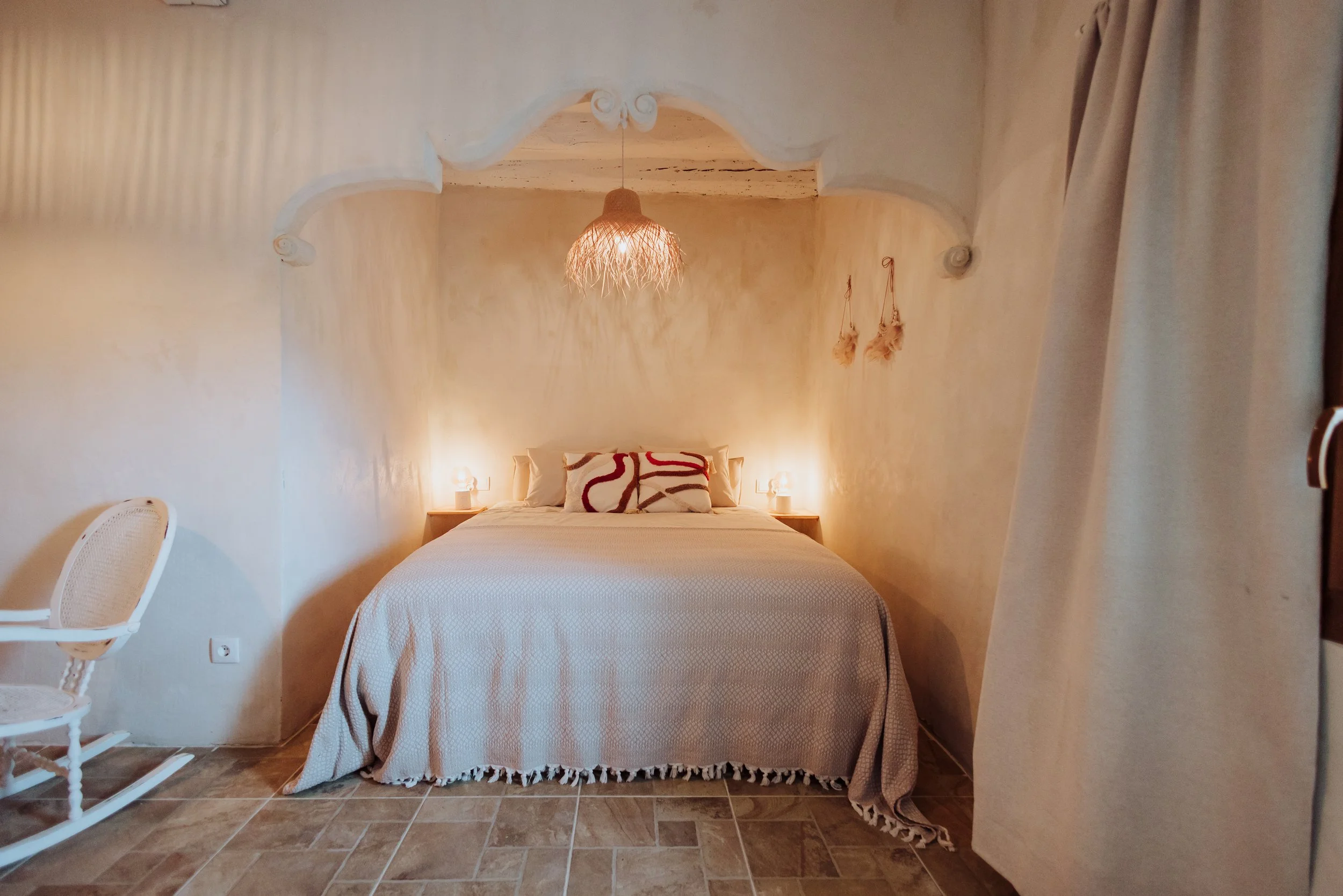 A cozy bedroom with a bed covered in a beige bedspread, featuring two decorative pillows with red and white patterns. The room has beige walls, a rustic hanging light fixture, and small bedside lamps. To the left, there is a white rocking chair, and to the right, a beige curtain is partially visible. The room has a warm, inviting ambiance.