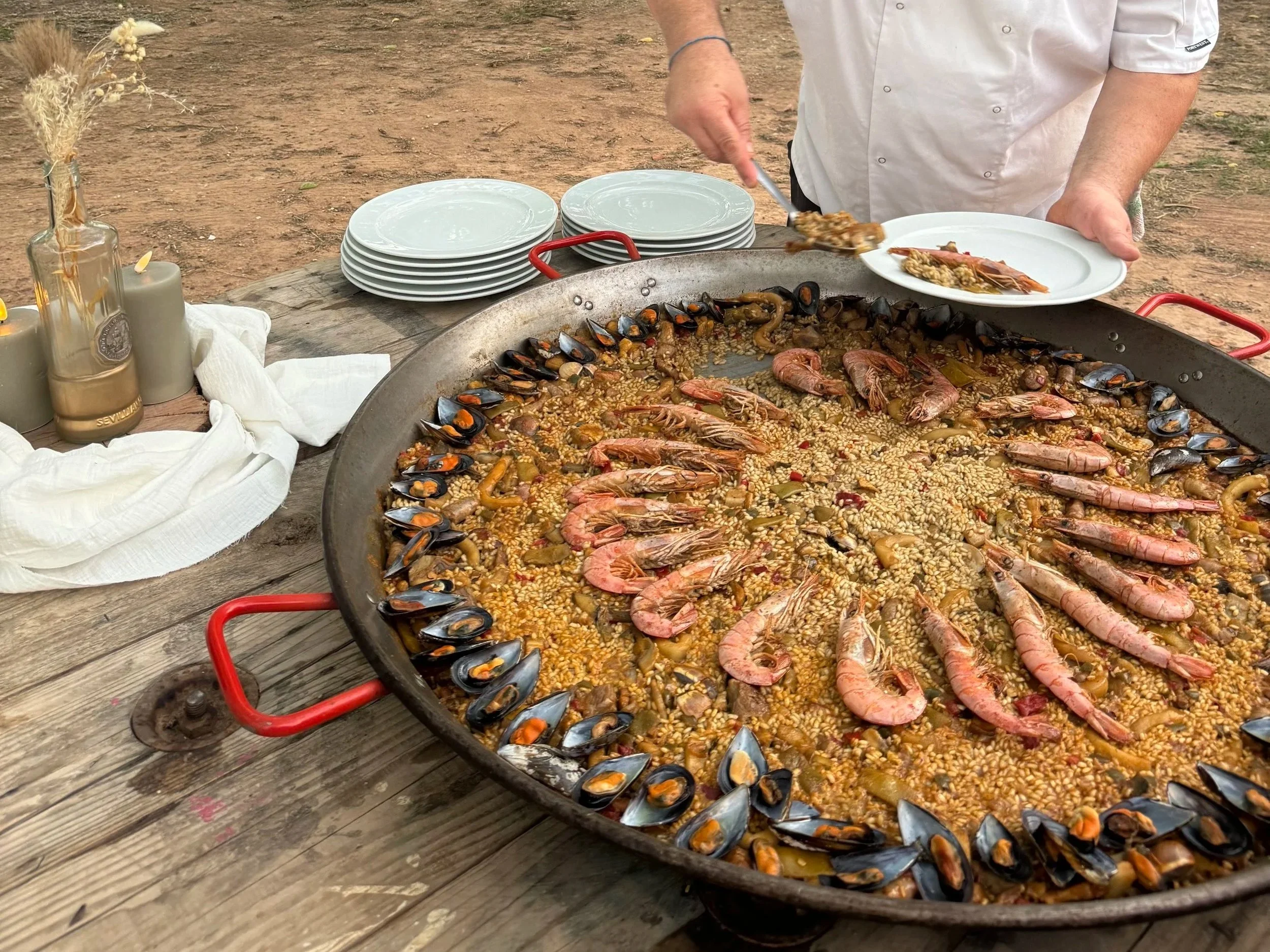 A large outdoor paella dish filled with rice, shrimp, mussels, and other seafood. A person in a white shirt is serving the paella onto a white plate.