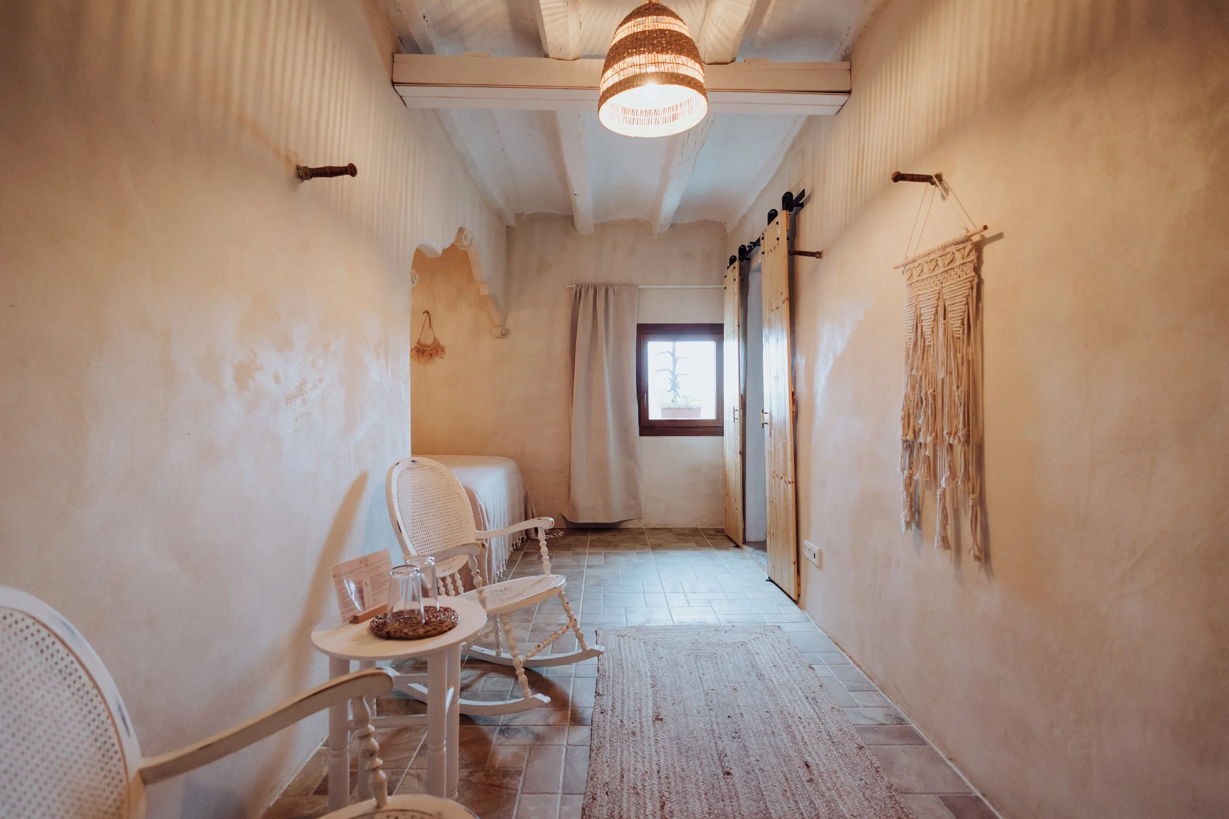 A cozy hallway with beige textured walls, wooden ceiling beams, and tile flooring. Wooden hooks are mounted on the walls, and there is a decorative woven wall hanging. Natural light enters through a small window, and chairs and a small table with glassware and a snack are present.