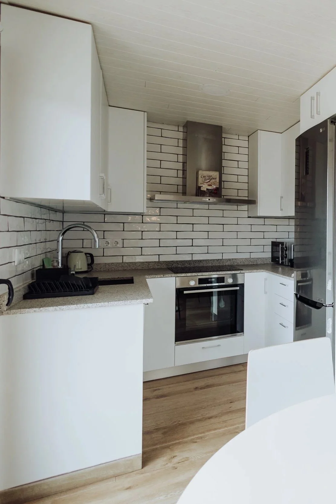 Modern kitchen with white cabinets, a speckled countertop, stainless steel oven, refrigerator, and a brick-style backsplash. There is a black dish rack, toaster, and a kettle on the counter.