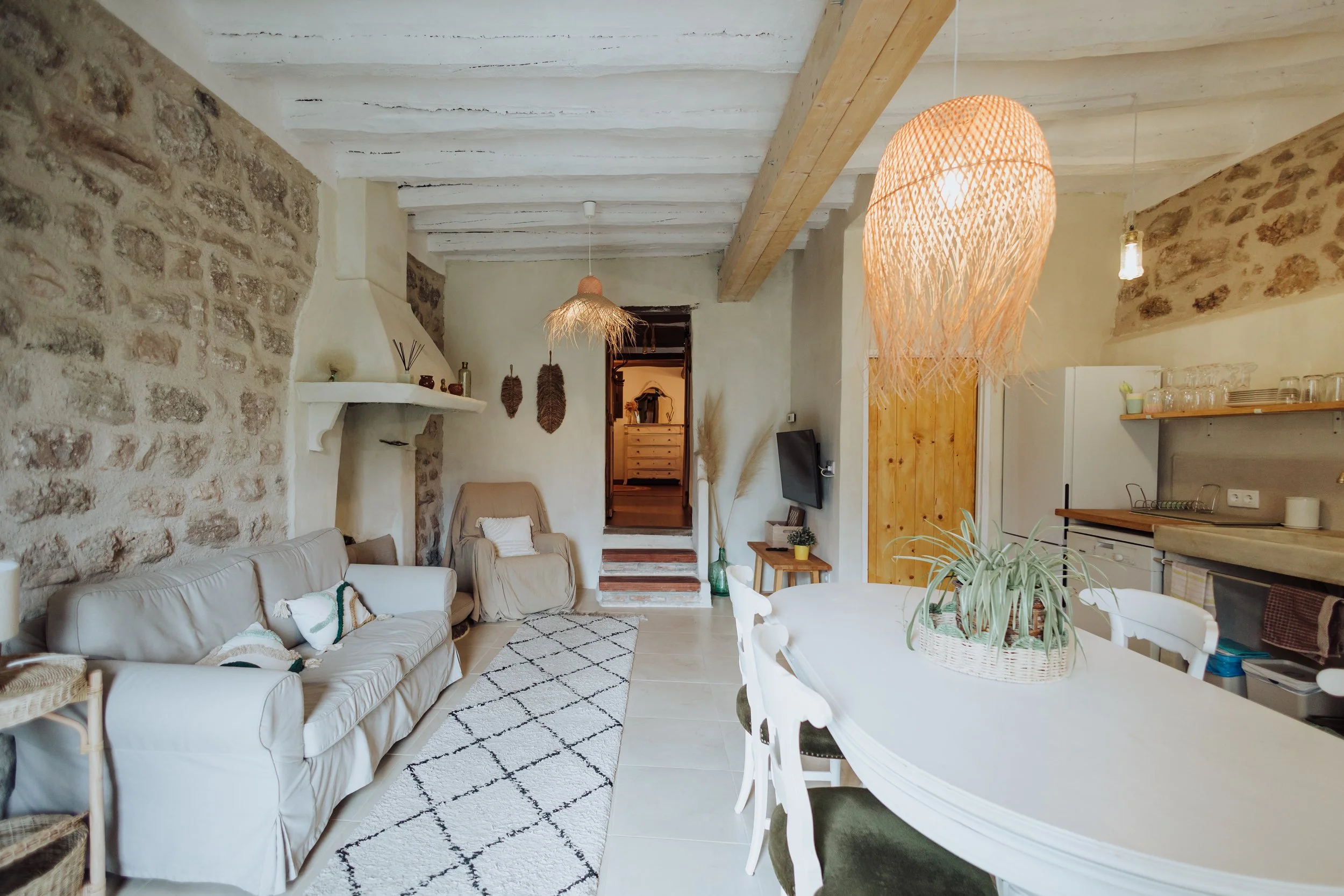 Living room with stone and white walls, white sofa with pillows, wooden beams and pendant lights, dining table with chairs, and open kitchen area with glassware.