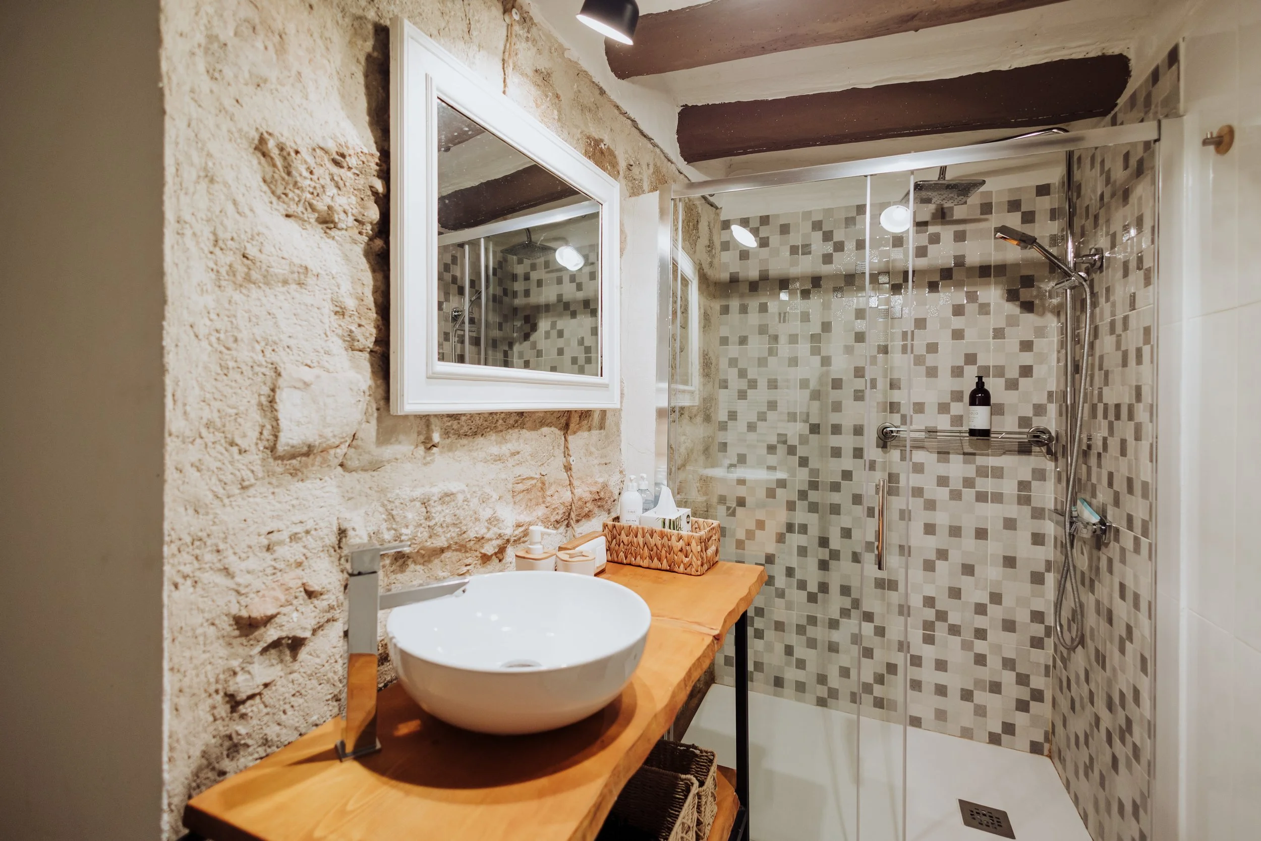 A rustic bathroom with a stone wall, a wooden countertop with a round vessel sink, a mirror, and a walk-in shower with mosaic tiles and a glass door.