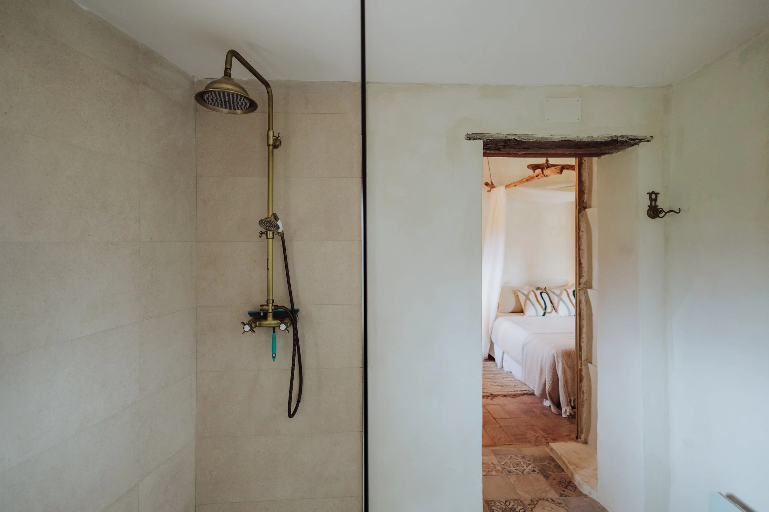 A bathroom with a showerhead and faucet on the left, separated from a bedroom by a wall opening. The bedroom is decorated with a bed, pillows, and a rustic wooden beam above the doorway.