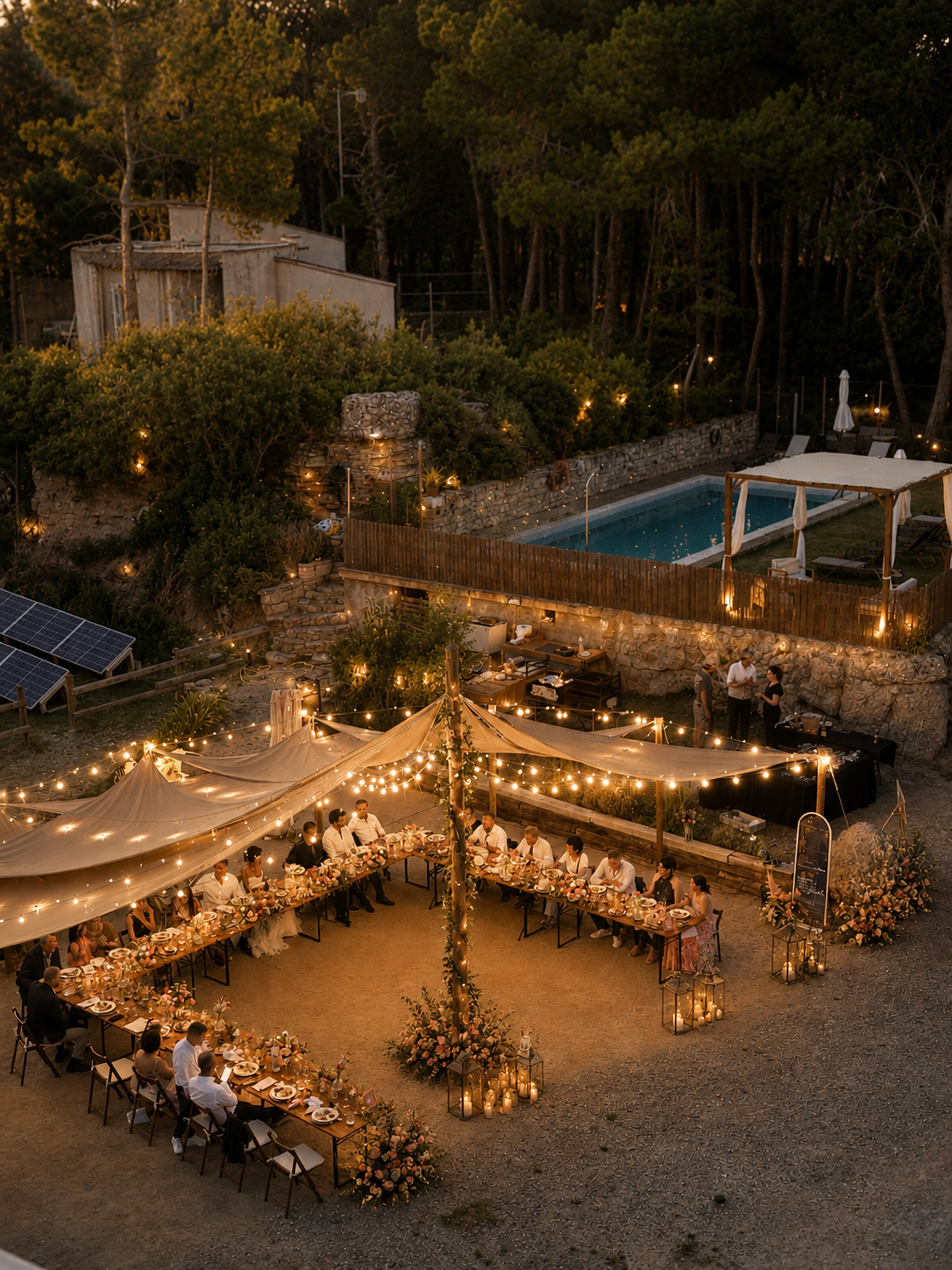 Outdoor wedding reception at dusk with long tables, string lights, floral centerpieces, and lanterns, set in a rustic yard with a pool in the background surrounded by trees.