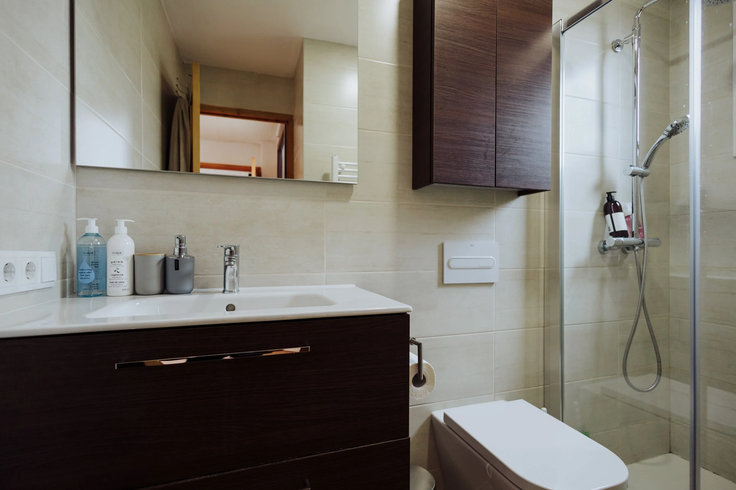 A modern bathroom with a beige tiled wall, a mirror above a dark wood vanity, and a shower area with a glass partition. The vanity has a white sink, soap and sanitizer bottles, and a soap dispenser. There is a toilet with a white lid and a wall-mounted flush panel. A mirror reflects the doorway and part of a room outside the bathroom.
