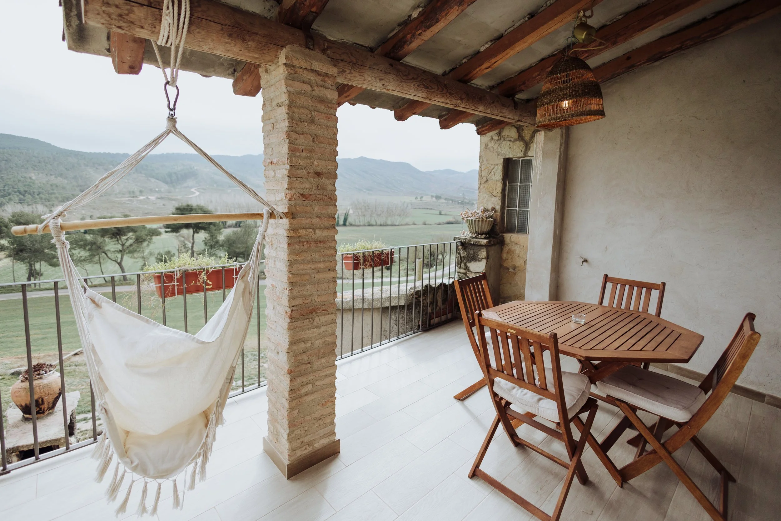 Balcony with wooden table and chairs, hanging hammock chair, potted plants, brick and stucco walls, and mountainous landscape view.