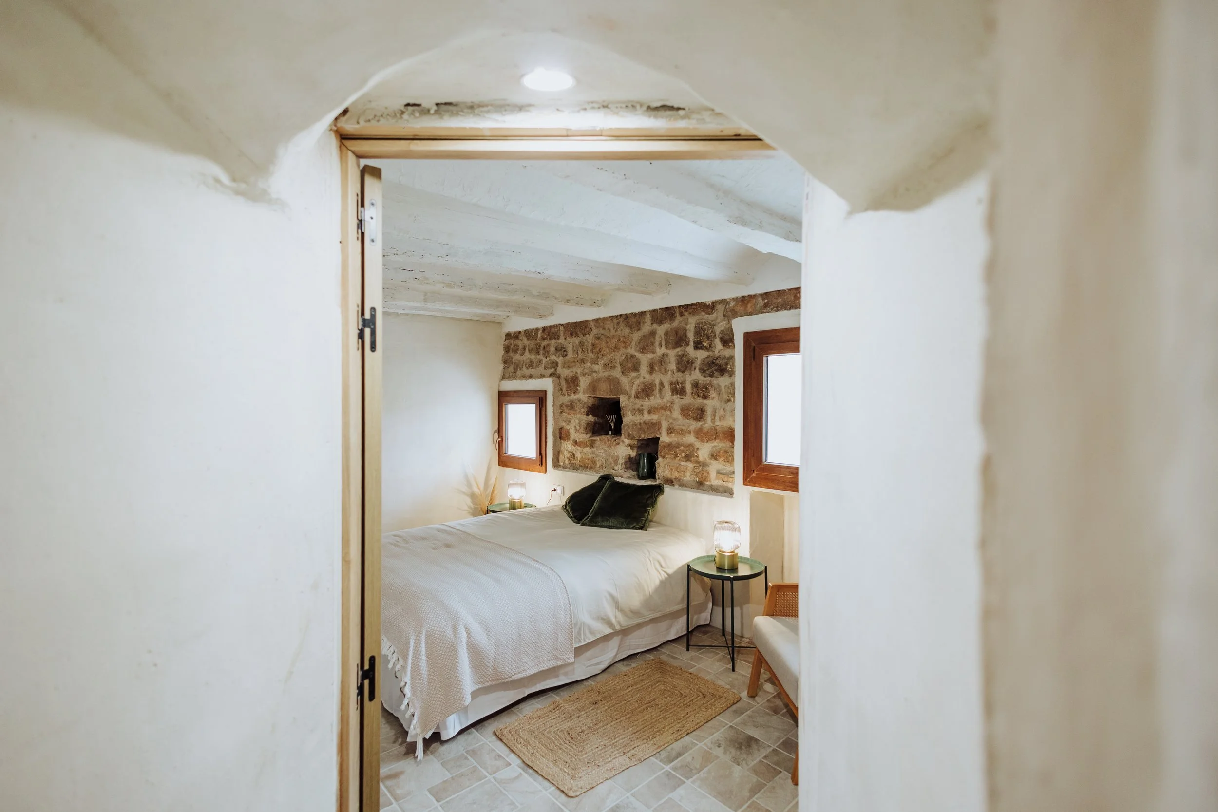 A cozy bedroom with a bed, two small side tables, and a rustic stone accent wall behind the headboard.