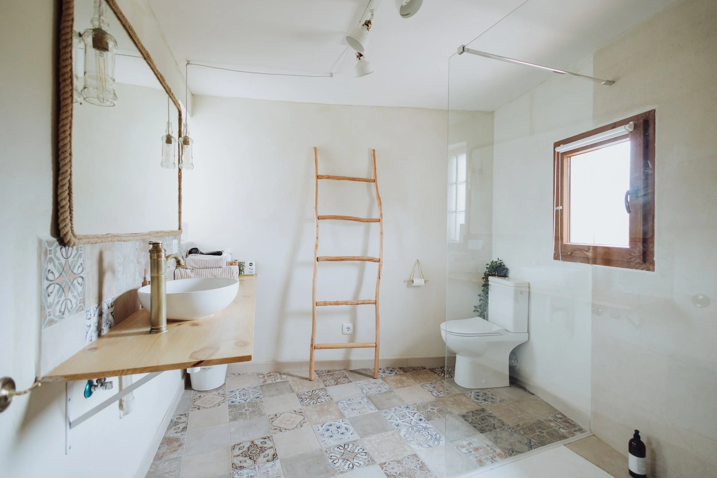 Bathroom with a wooden ladder, a window with blinds, a toilet, a white sink with a gold faucet, a mirror, and decorative tile flooring.